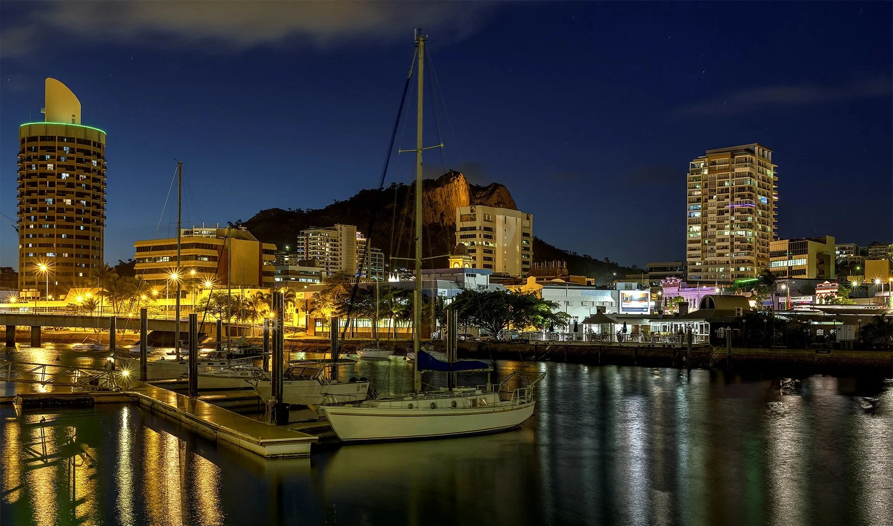 A yacht is moored at a marina at night with illuminated buildings and a hill in the background under a clear sky.