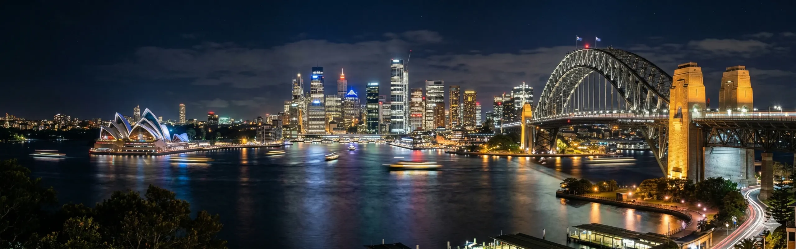 Sydney skyline at night with illuminated buildings, the Sydney Opera House on the left, and Sydney Harbour Bridge spanning the harbour on the right.