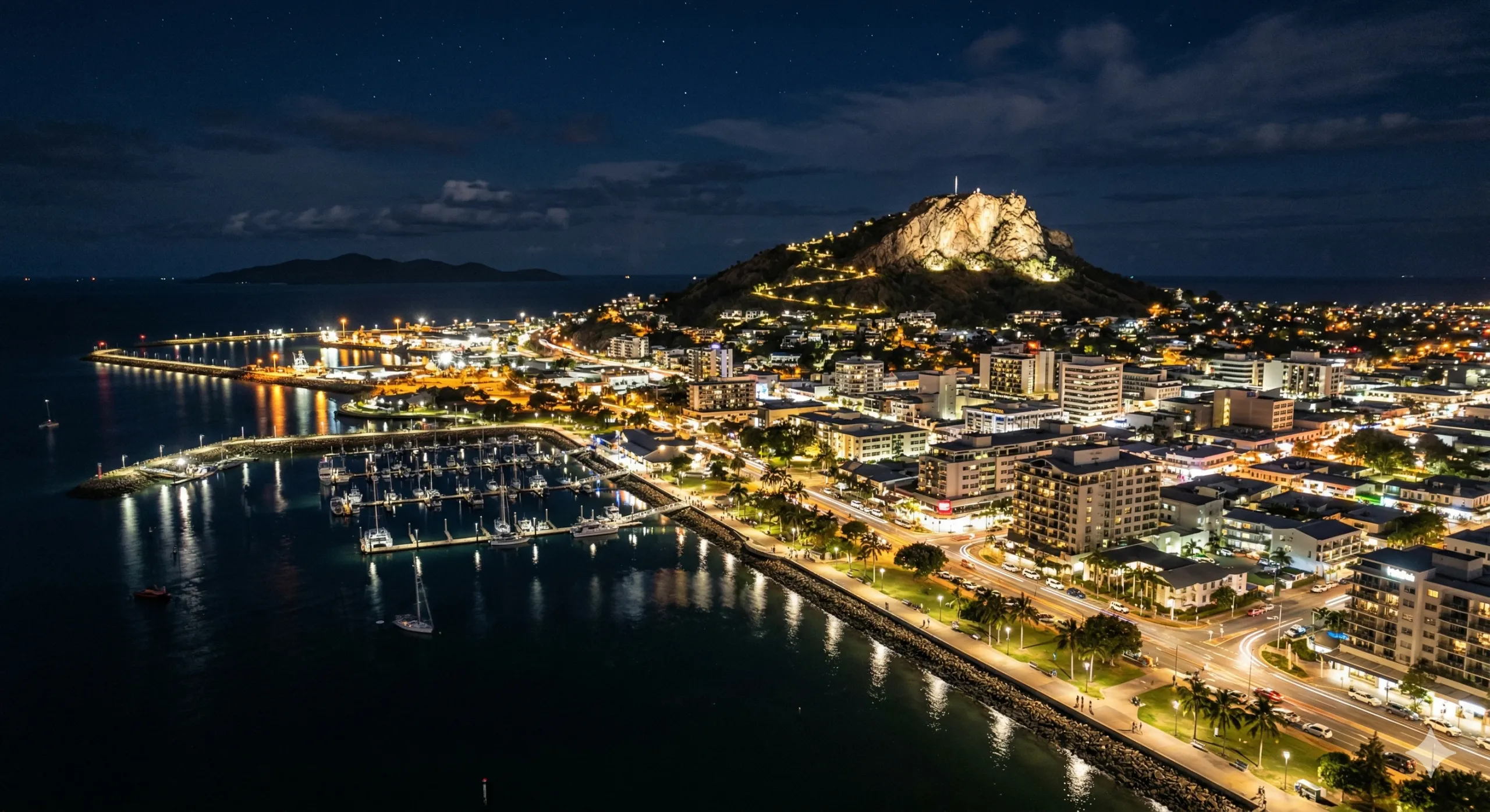 Aerial view of a coastal city at night with illuminated buildings, a marina, and a hill with a lit-up lookout point in the background.