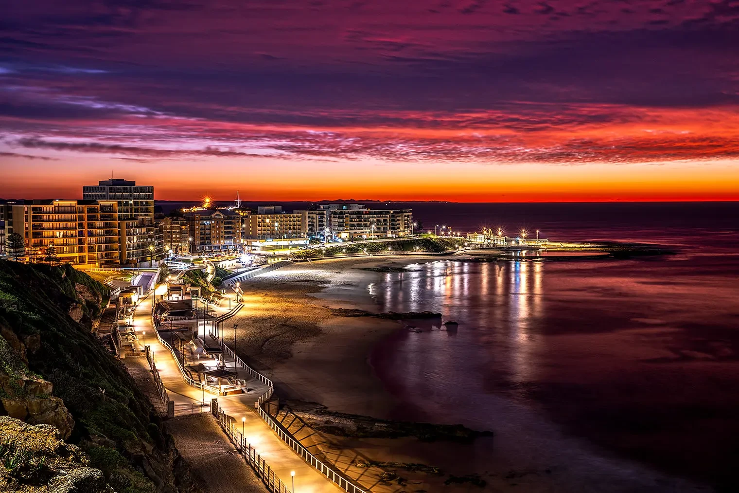 A coastal cityscape at dusk features lit walkways, modern buildings, a sandy beach, and a vibrant purple and orange sunset sky reflecting on calm water.