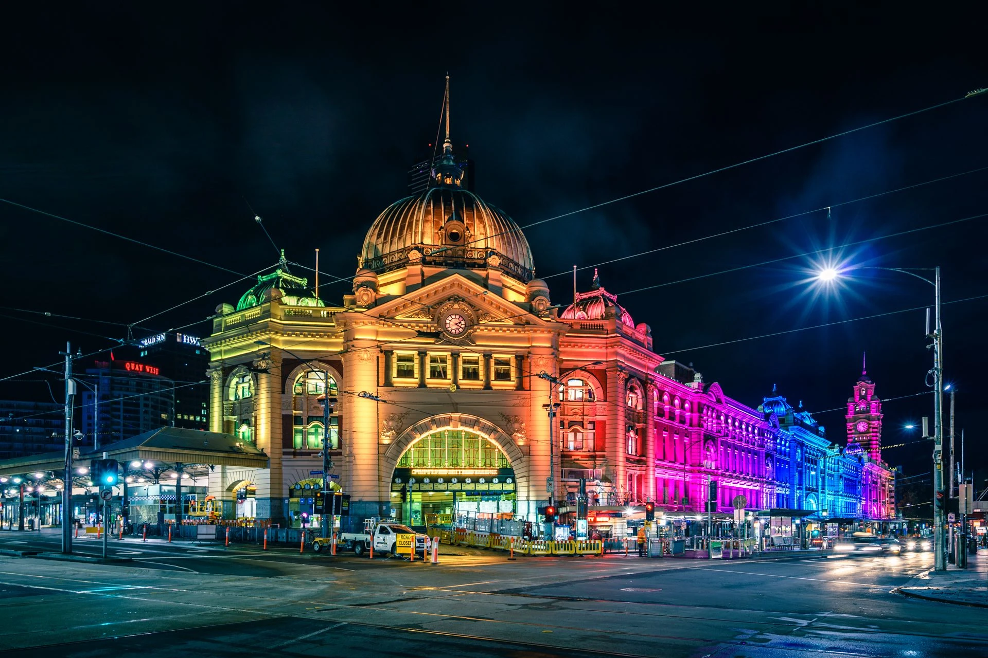 Historic railway station building illuminated with vibrant rainbow-coloured lights at night, with tram wires and city street in the foreground.