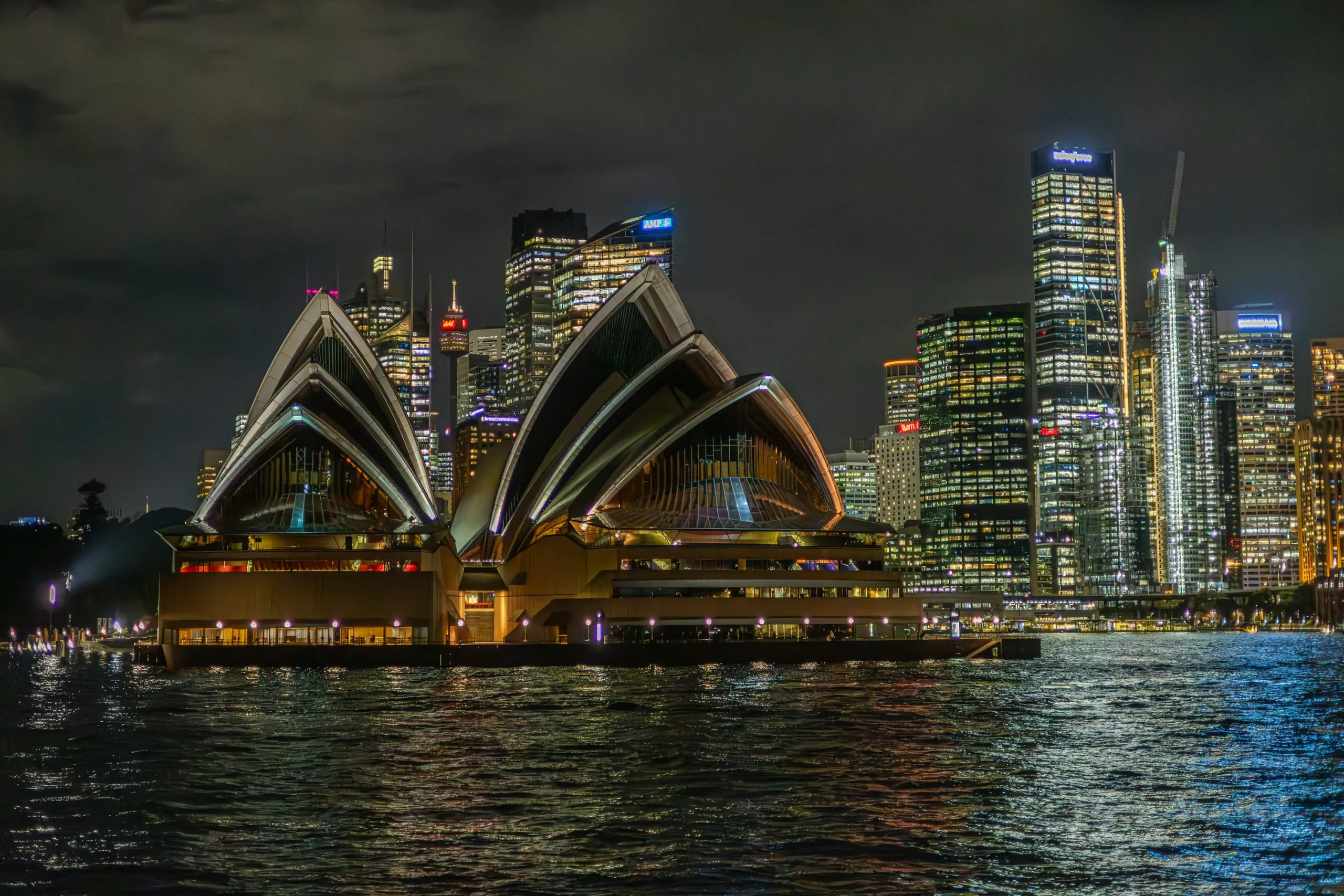 The Sydney Opera House illuminated at night, with city skyscrapers lit up in the background and reflections visible on the harbour water.