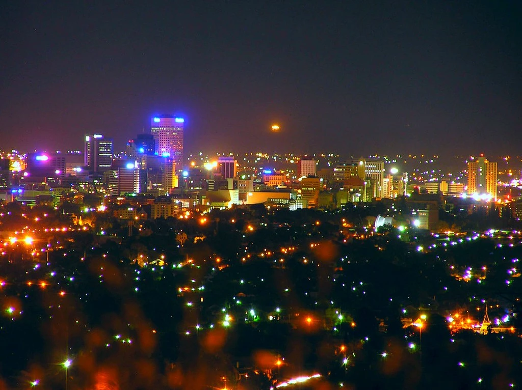A city skyline at night with illuminated buildings and colourful lights, set against a dark sky with a bright glow on the horizon.