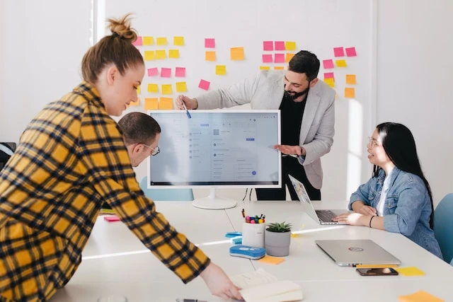 Four people in a modern office collaborate around a desk with a computer monitor, sticky notes on the wall, and various office supplies.