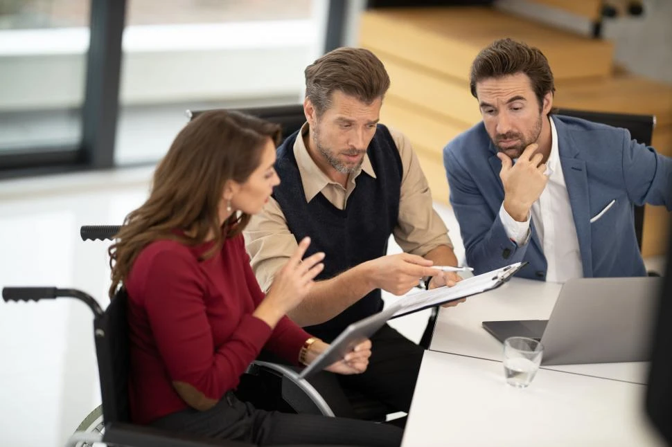 Three people sit at a table in discussion, looking at documents and a laptop, with one person in a wheelchair leading the conversation.