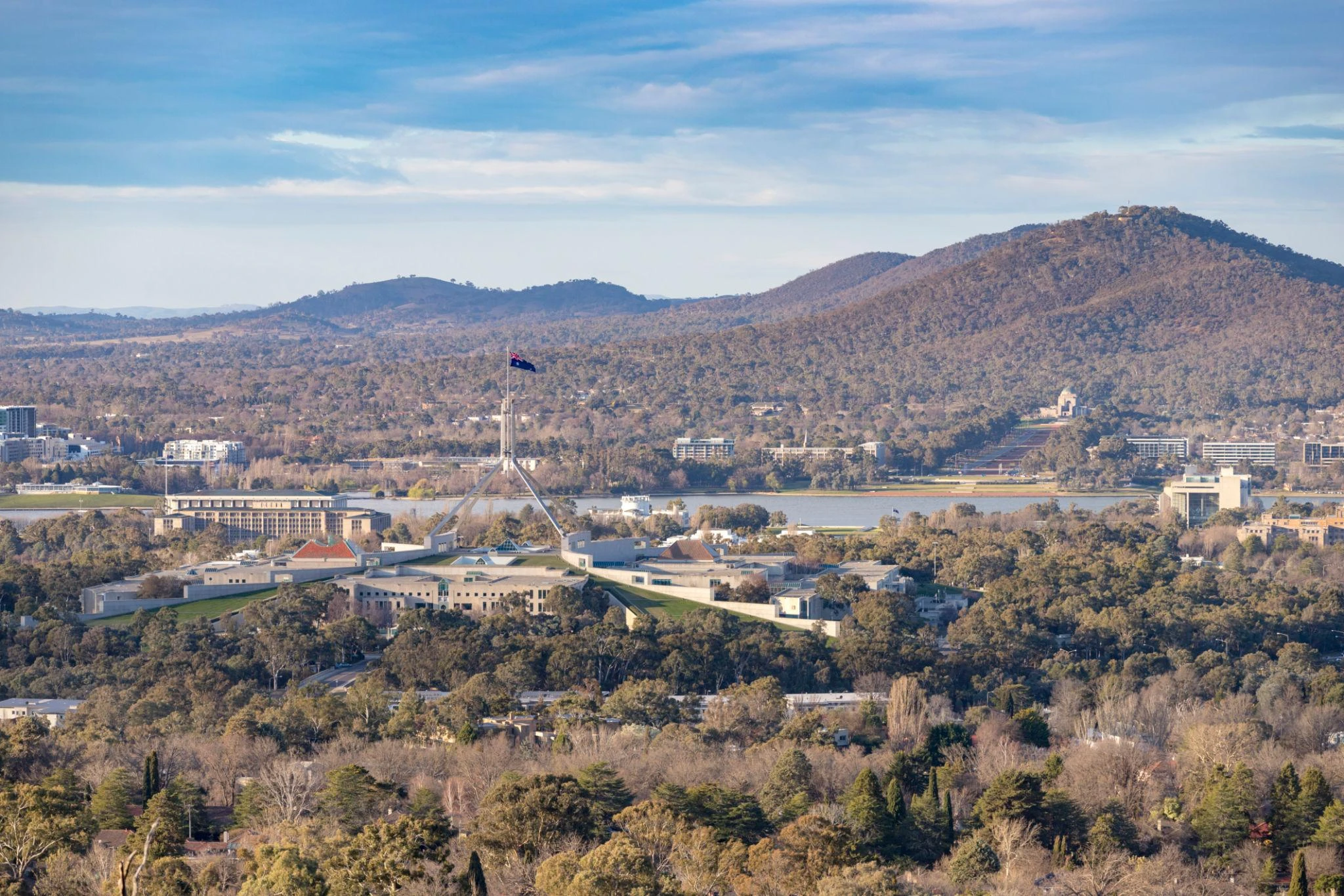 Aerial view of a government building with a large flagpole, surrounded by trees and hills under a partly cloudy sky.