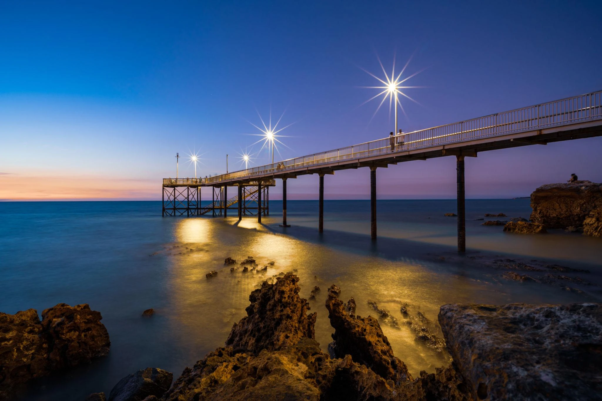 A long pier with lit street lamps extends over calm water at dusk, with rocks visible in the foreground and a clear sky in the background.