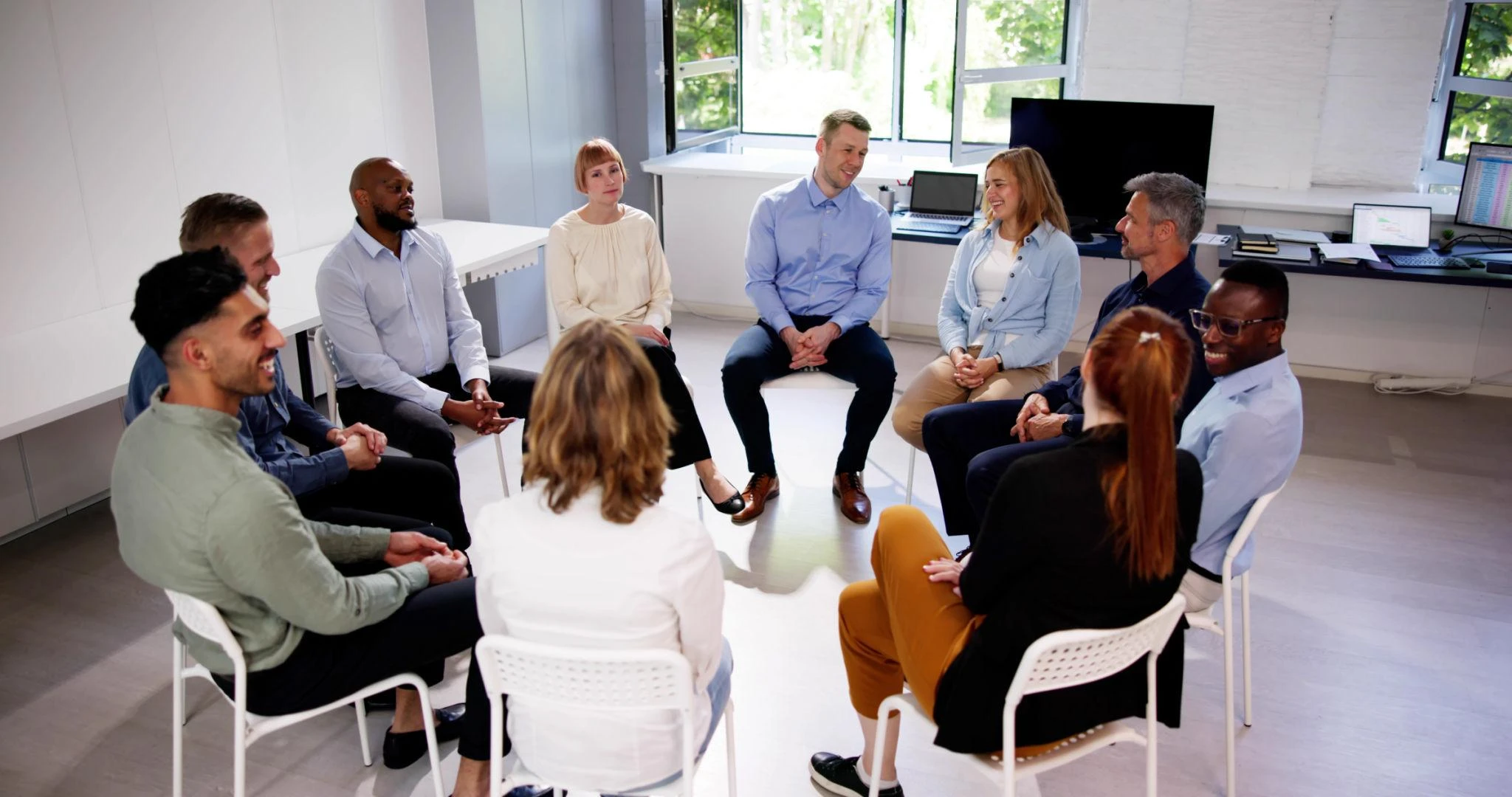 A group of people sit in a circle on chairs in a bright room, engaged in discussion or a group meeting.