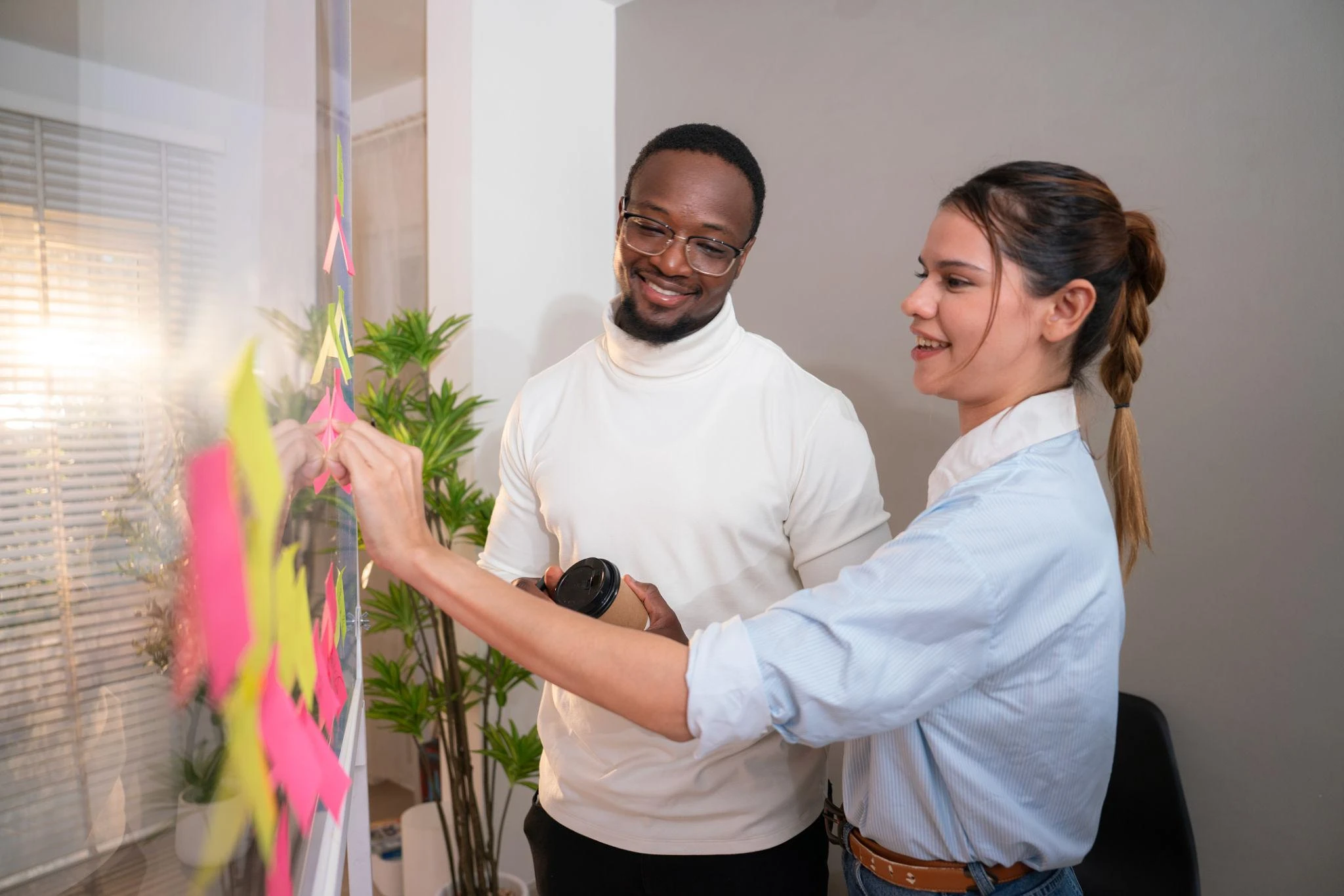 Two colleagues stand by a glass board with colourful sticky notes, collaborating and discussing ideas in an office setting.