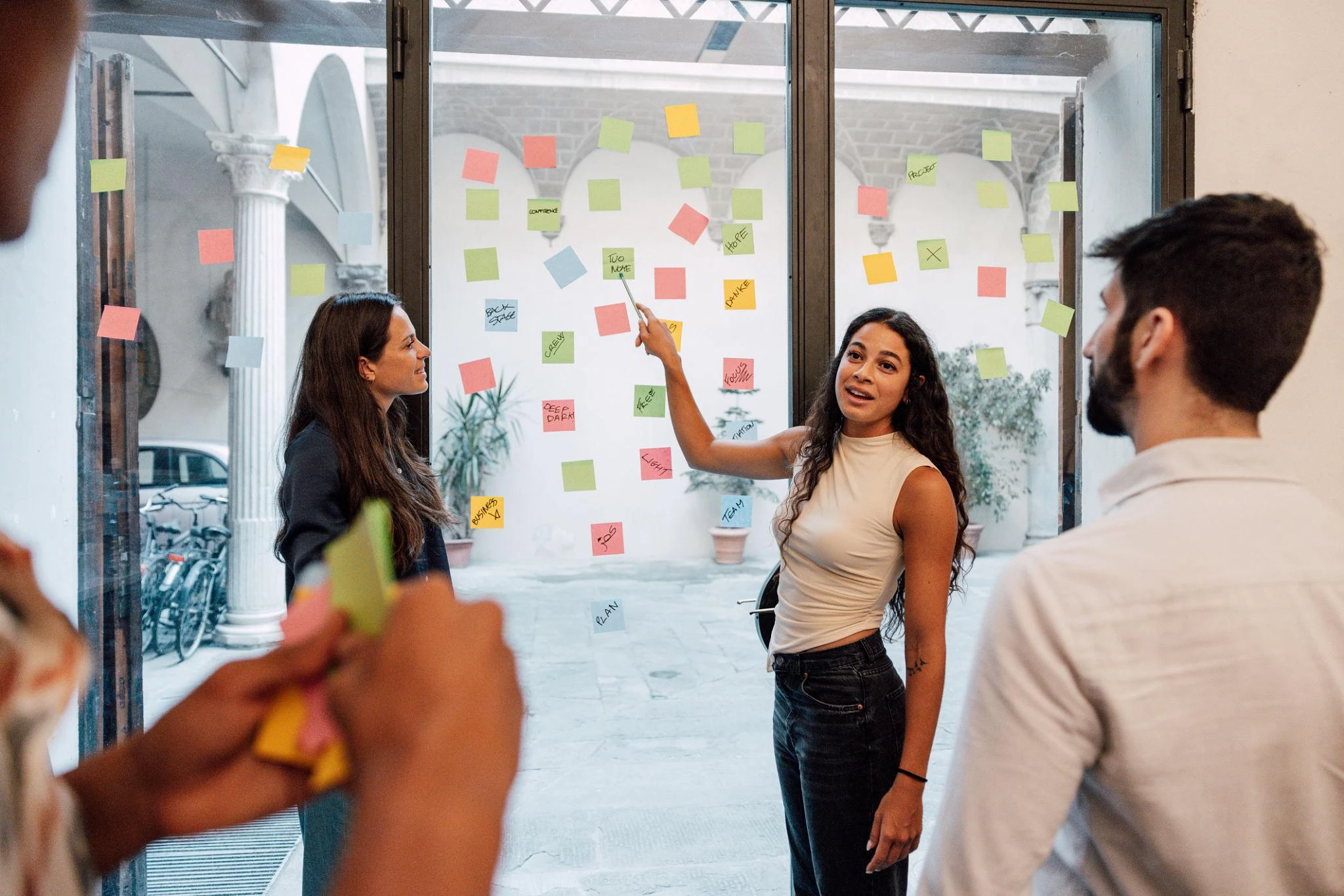 Four people stand by glass doors covered with colourful sticky notes, discussing and brainstorming ideas together. One woman gestures towards the notes whilst speaking.