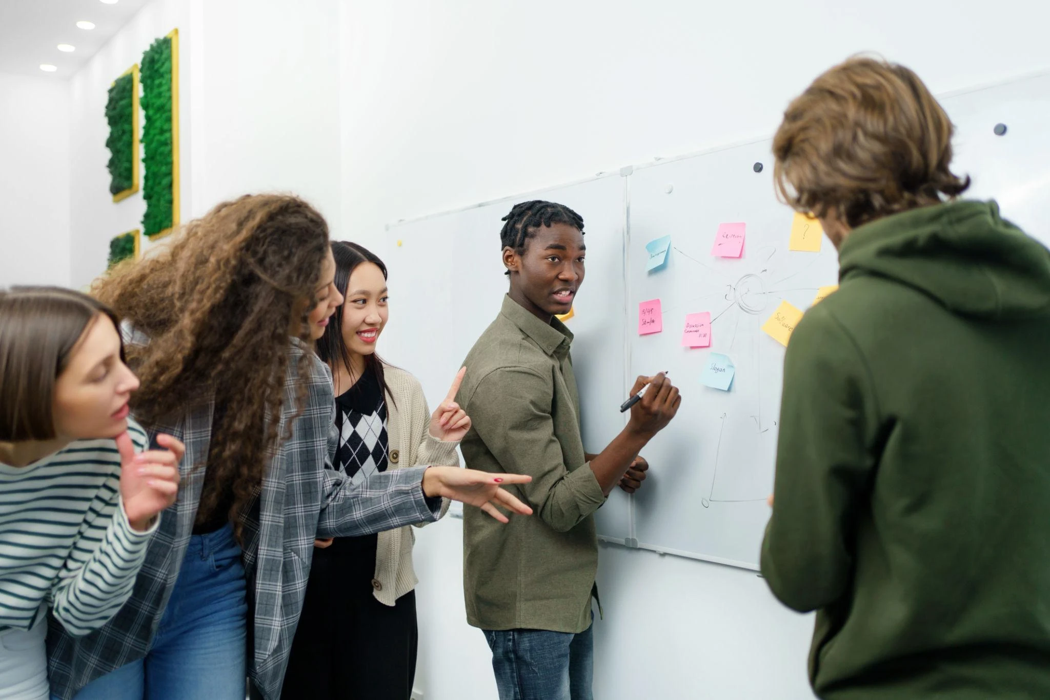 Five young adults collaborate at a whiteboard covered with sticky notes, whilst one person writes and others engage in discussion.