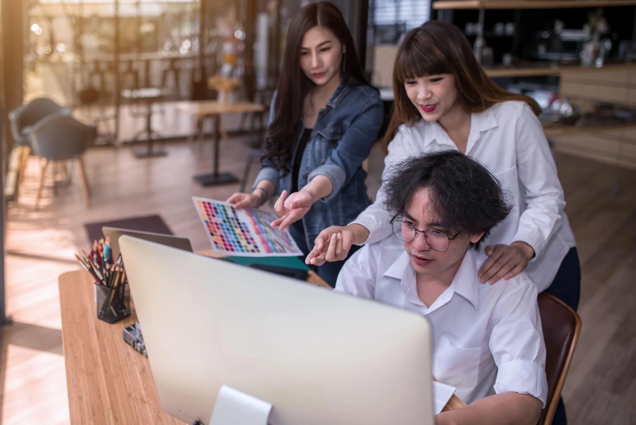 Two women and one man working together at a desk; the man is using a computer while the women point at the screen and hold a colour swatch book.