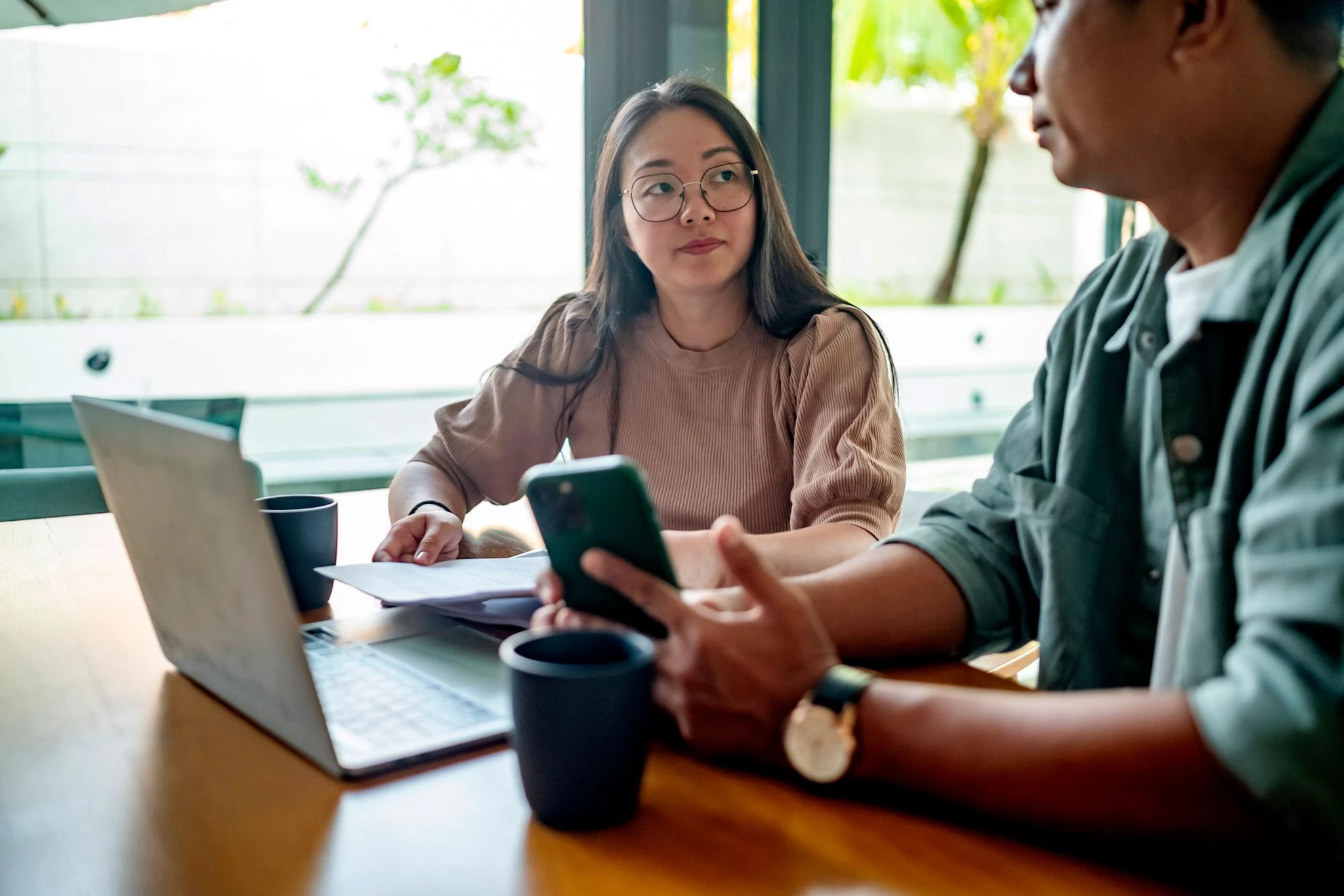 Two people sit at a table with a laptop, mobile phone, papers, and coffee cups, having a discussion in a bright indoor space with large windows.