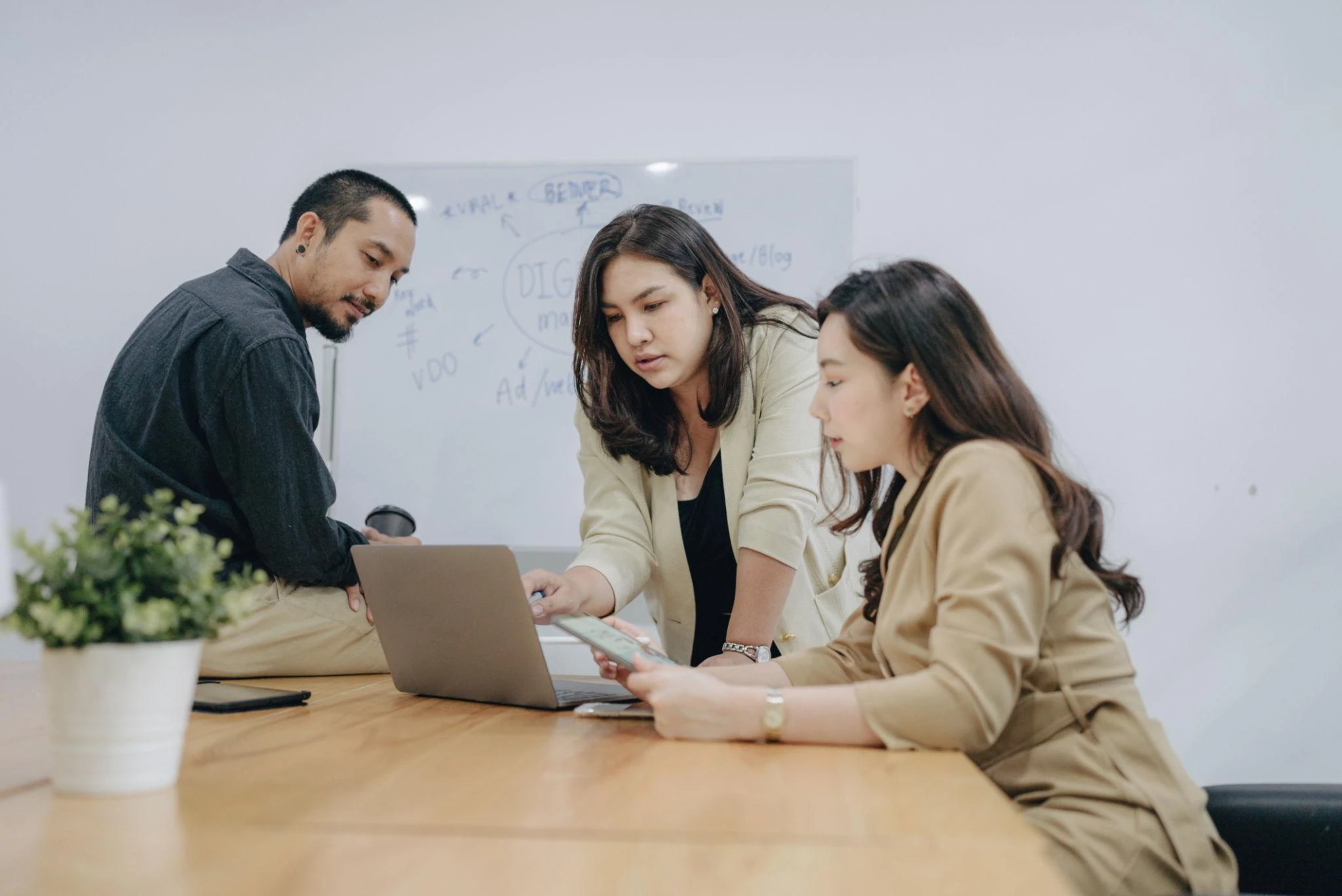 Three people in business attire discuss work at a table with a laptop and documents, with a whiteboard in the background.