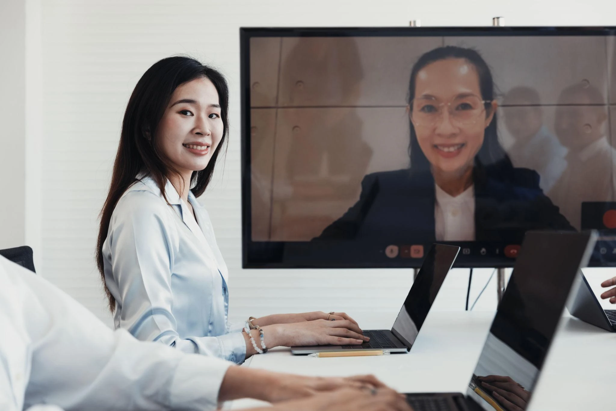 A woman sits at a conference table with a laptop, smiling, whilst another woman participates via video call on a large screen in the background.