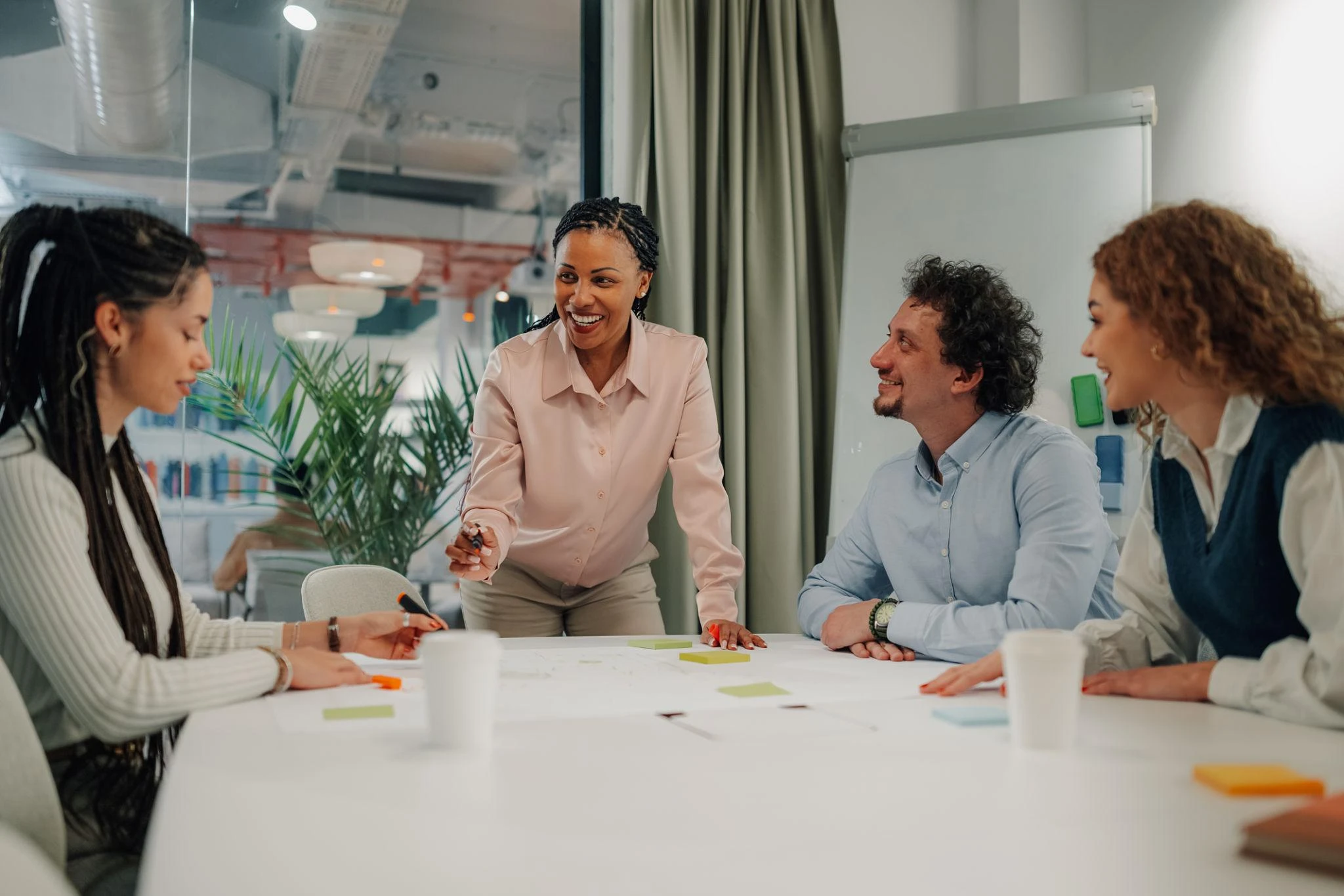 Four people sit and stand around a table in an office, discussing documents and sticky notes, with one woman standing and leading the discussion.