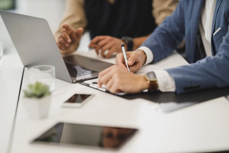 Two people sit at a desk with a laptop, tablet, mobile phone, and documents; one person writes whilst the other gestures during a discussion.