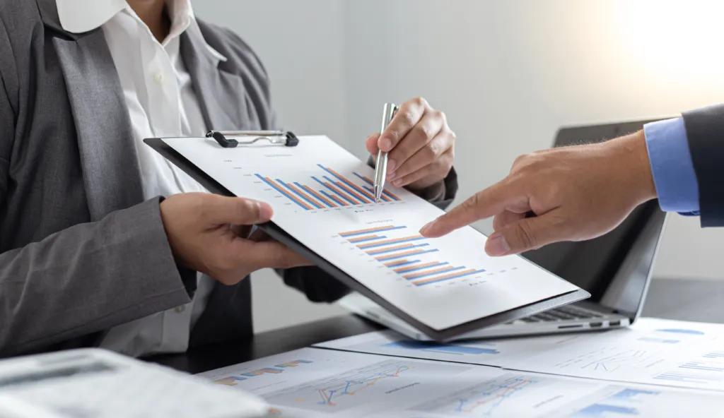 Two people in business attire review bar graphs and charts on a clipboard, discussing data analysis with documents and a laptop on the table.