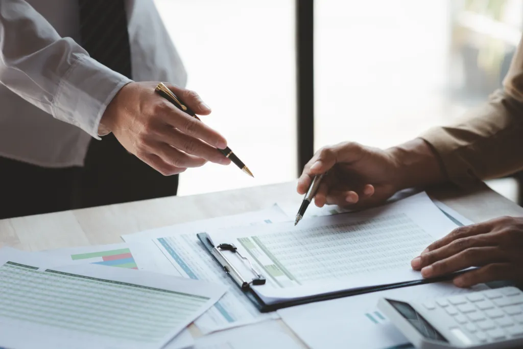 Two people review and discuss documents with charts and data at a desk, each holding a pen, with financial reports and graphs visible.