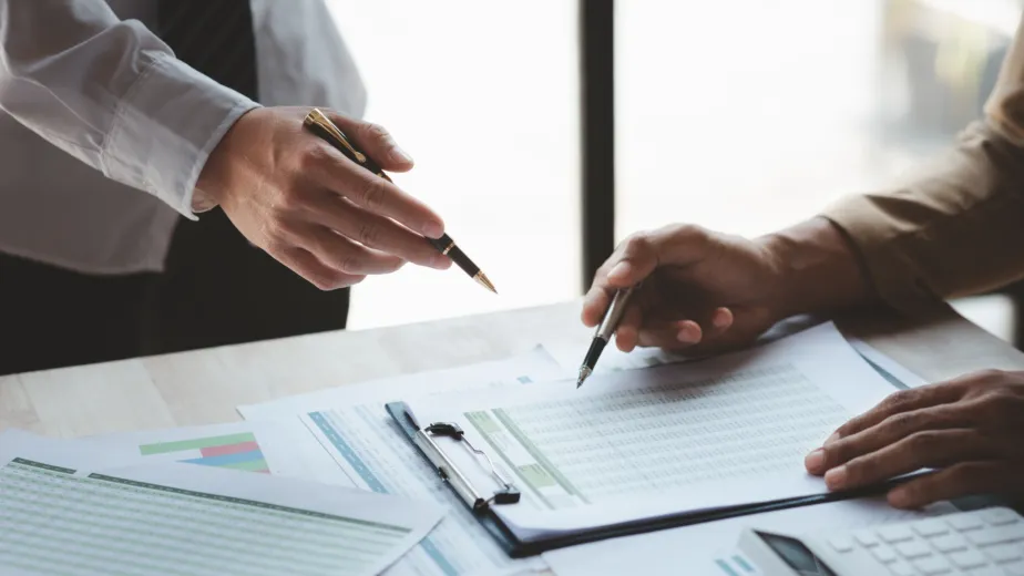 Two people review and discuss documents with charts and data at a desk, each holding a pen, with financial reports and graphs visible.