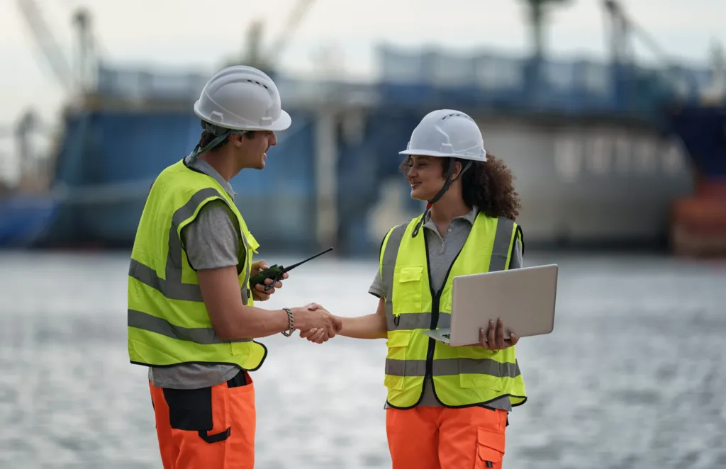 Two workers in safety vests and helmets shake hands near a waterfront; one holds a laptop and the other holds a walkie-talkie.