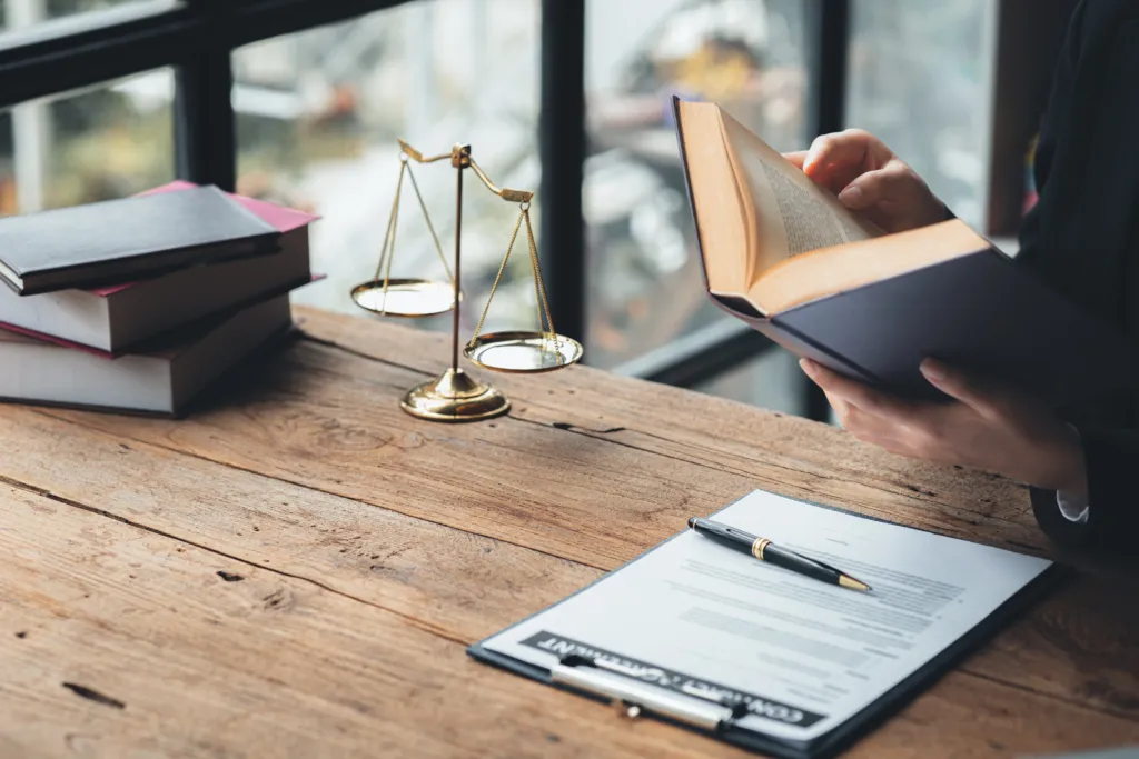 A person reads a book at a wooden desk with legal documents, a pen, stacked books, and a set of balance scales.