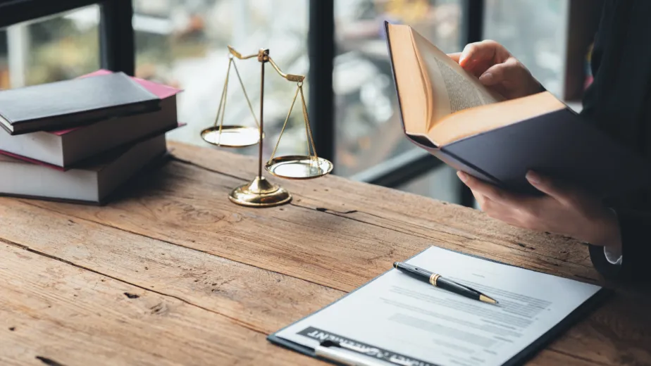 A person reads a book at a wooden desk with legal documents, a pen, stacked books, and a set of balance scales.