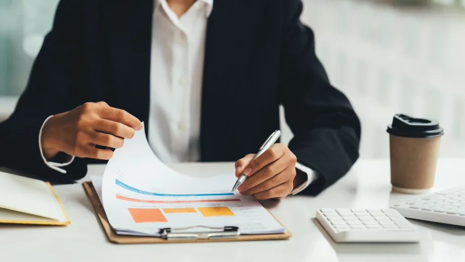 Person in business attire reviews documents with charts and graphs at a desk, holding a pen. A calculator, coffee cup, and folder are also on the desk.