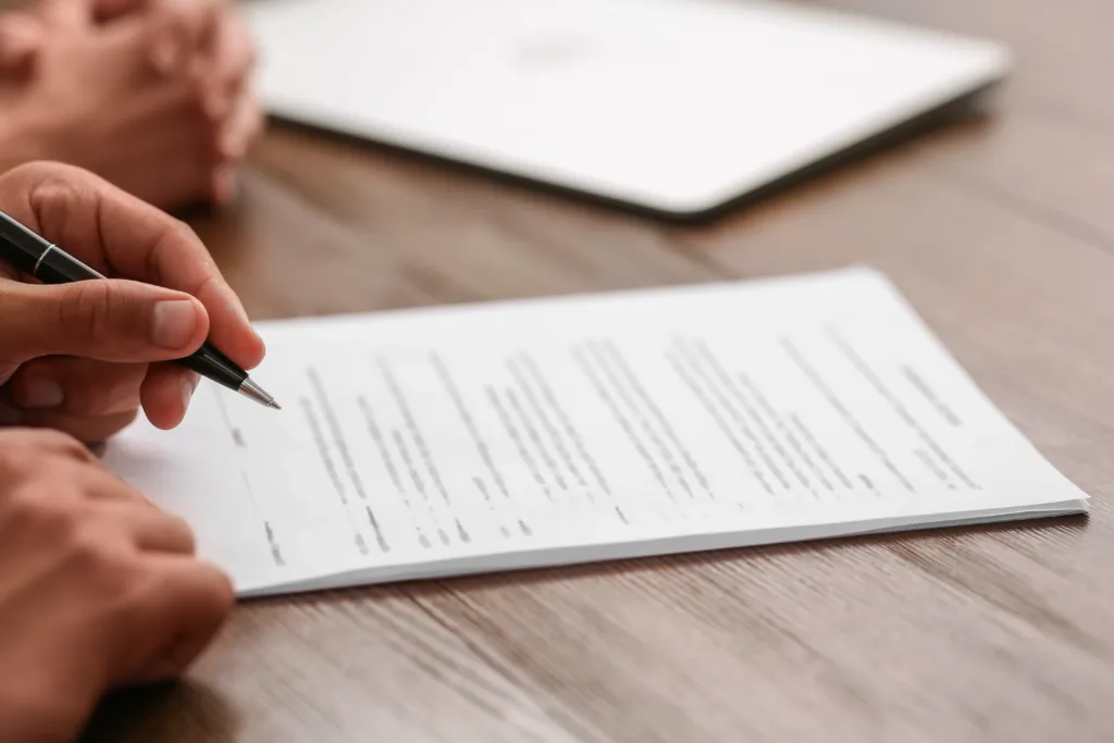 A person holds a pen and reviews a printed document on a wooden table, with another person’s hands and a closed laptop in the background.