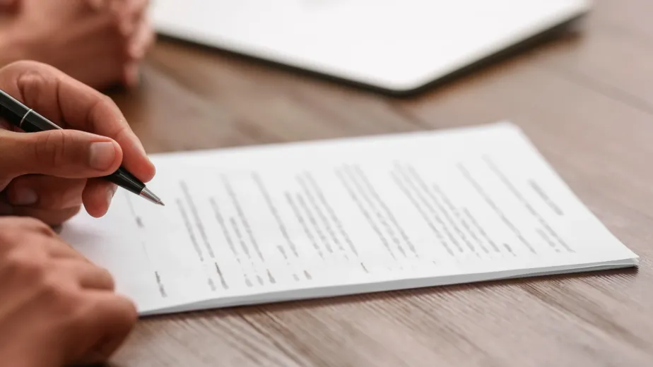 A person holds a pen and reviews a printed document on a wooden table, with another person’s hands and a closed laptop in the background.