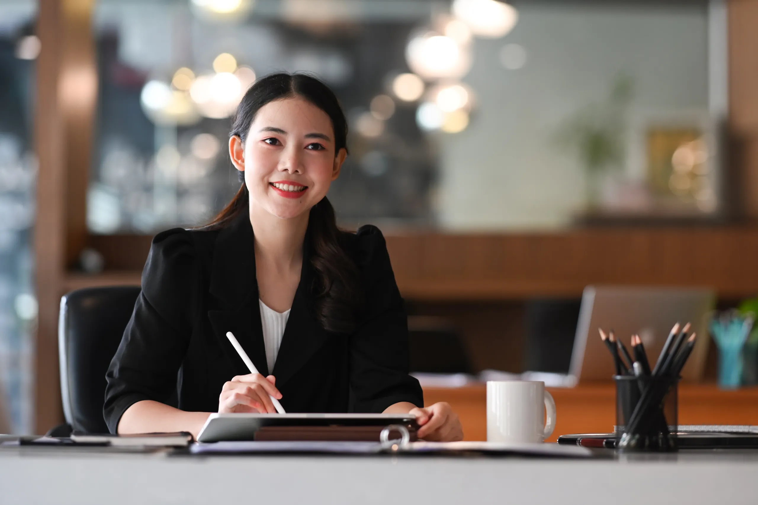 A woman sitting at a desk, smiling, holding a pen over a tablet. A white mug, pencil holder, and office supplies are on the desk. The background shows a blurred office setting.