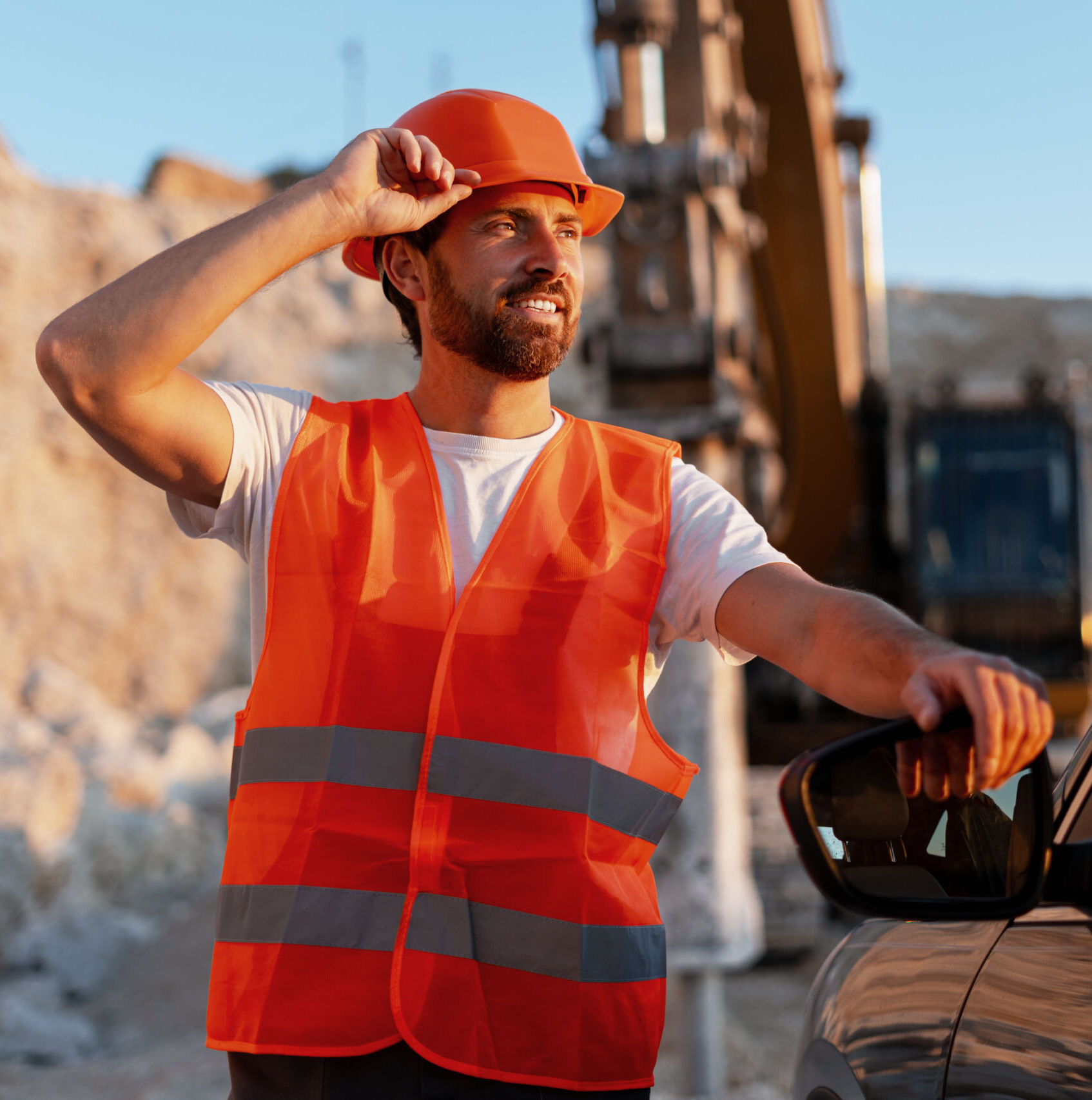 Man wearing an orange safety vest and hard hat stands next to a car at a construction site, with heavy machinery and soil in the background.