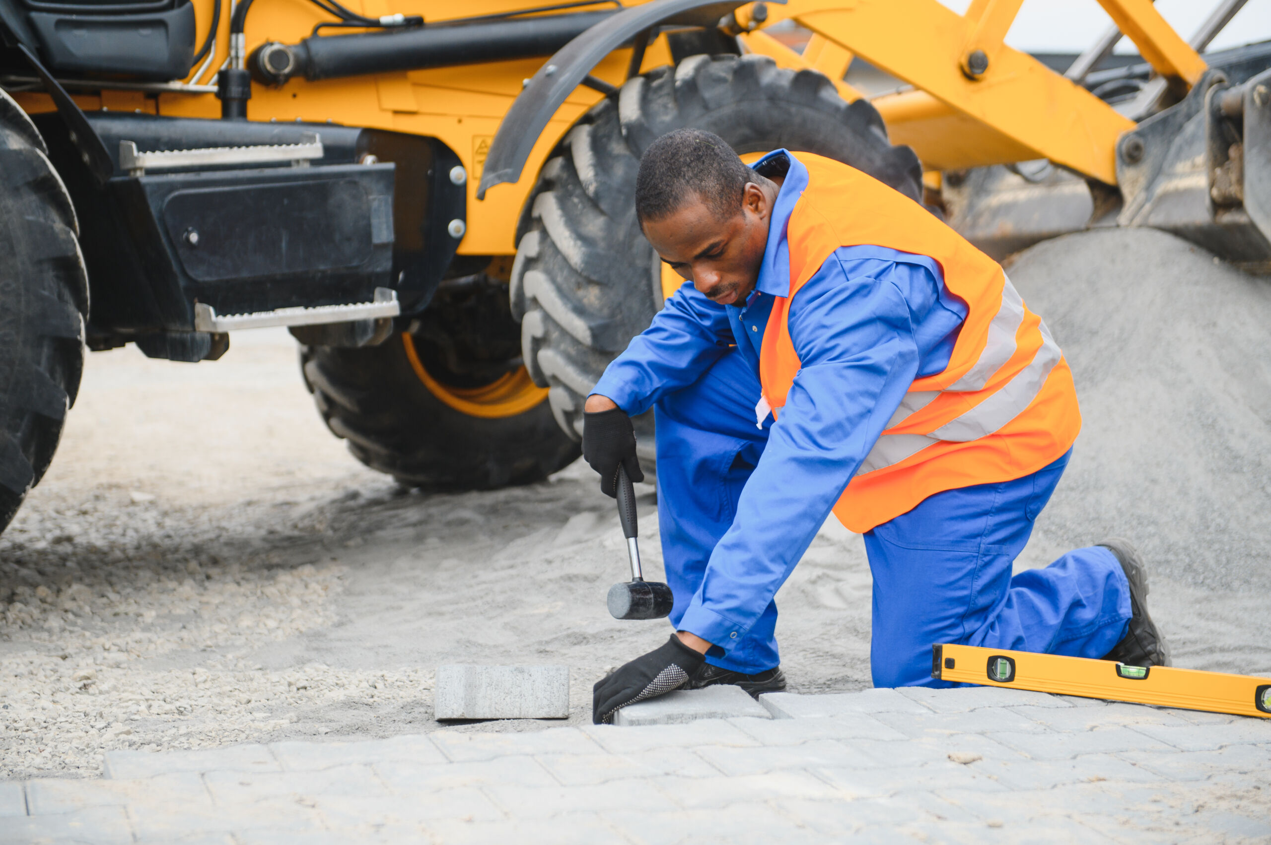 A construction worker in a blue uniform and orange safety vest uses a mallet to lay paving stones next to a yellow construction vehicle.