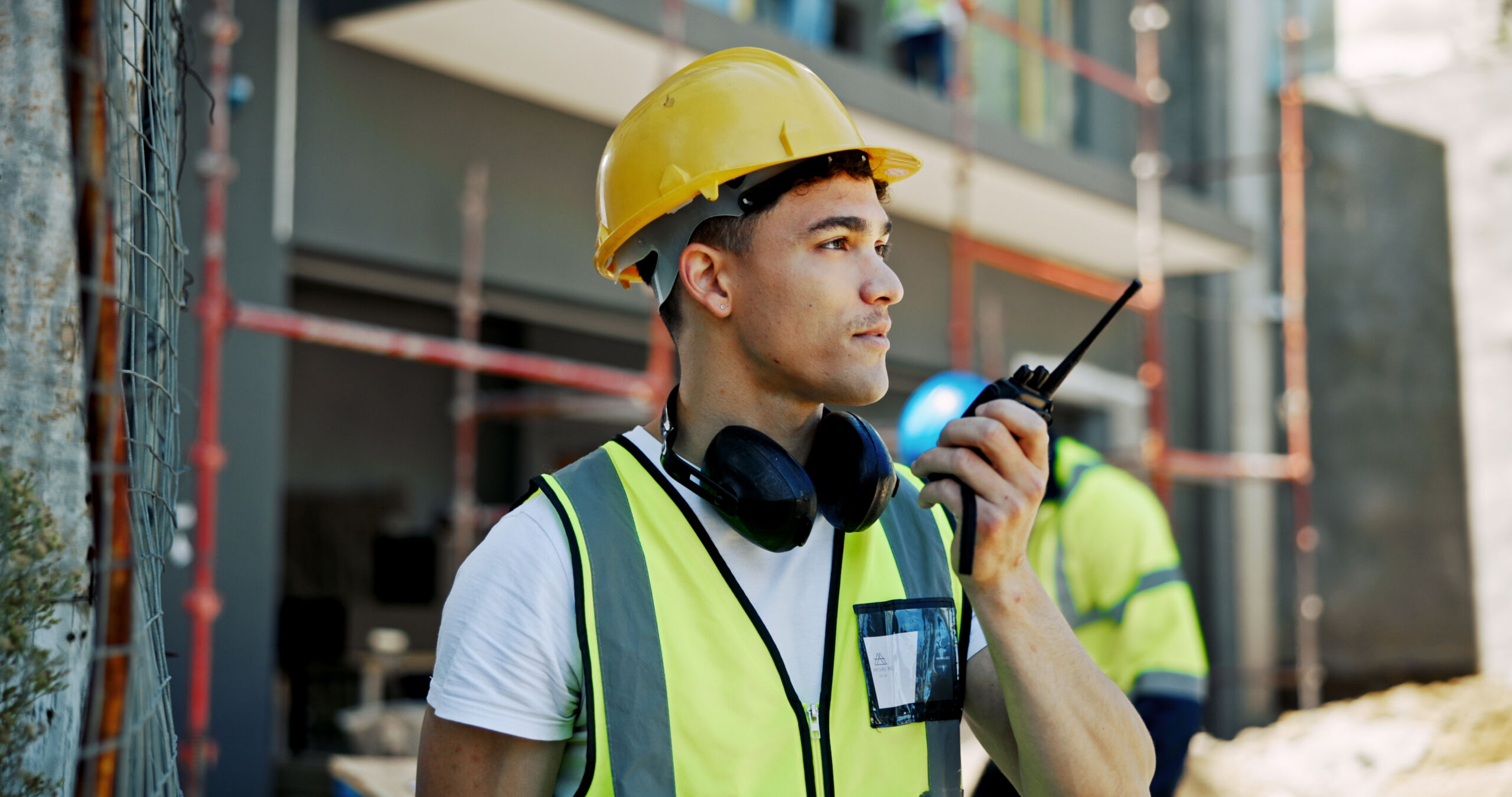 A construction worker wearing a yellow hard hat and safety vest communicates using a two-way radio at a building site.