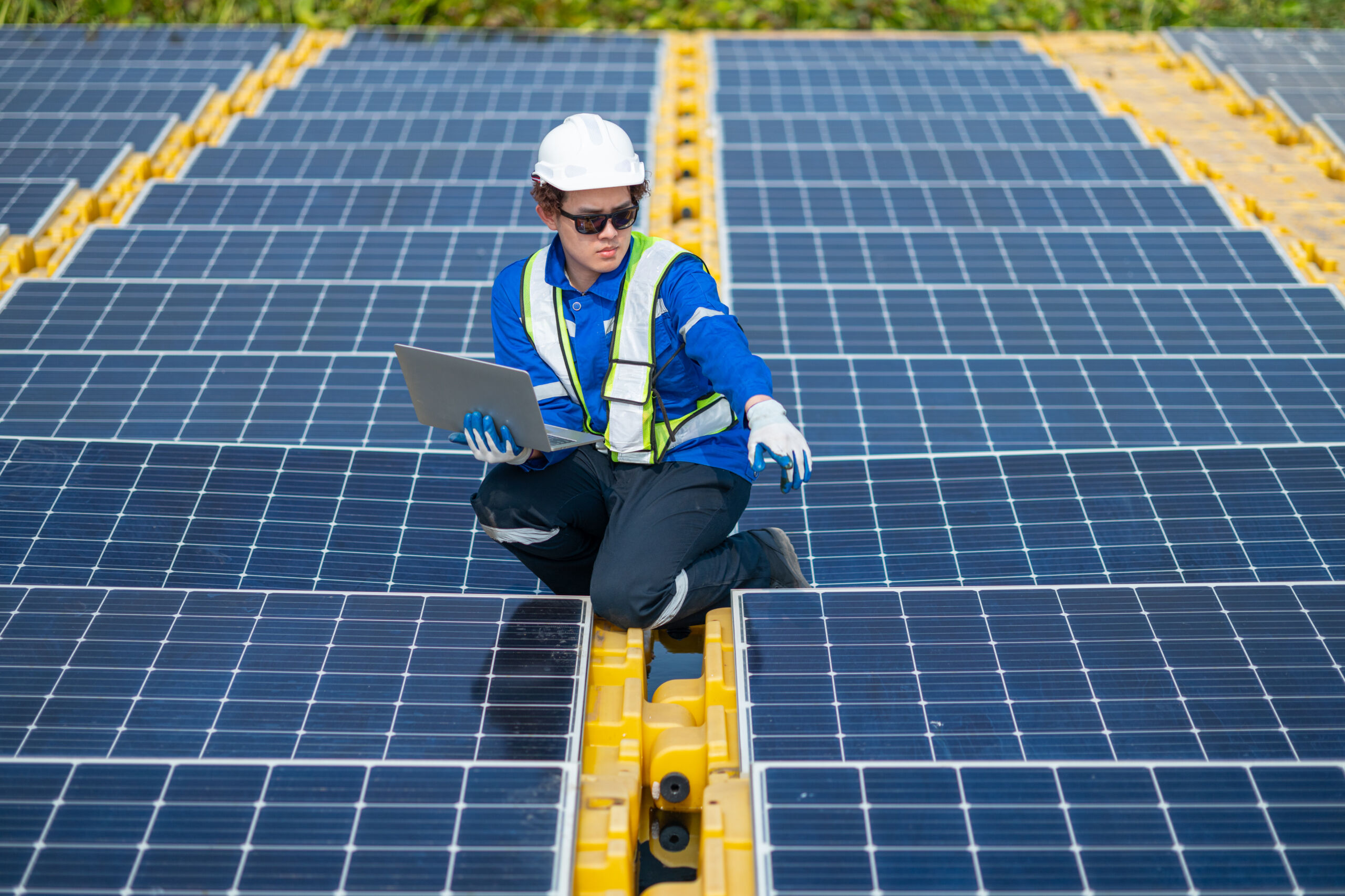 A worker wearing safety gear inspects solar panels whilst holding a laptop at a solar energy facility.