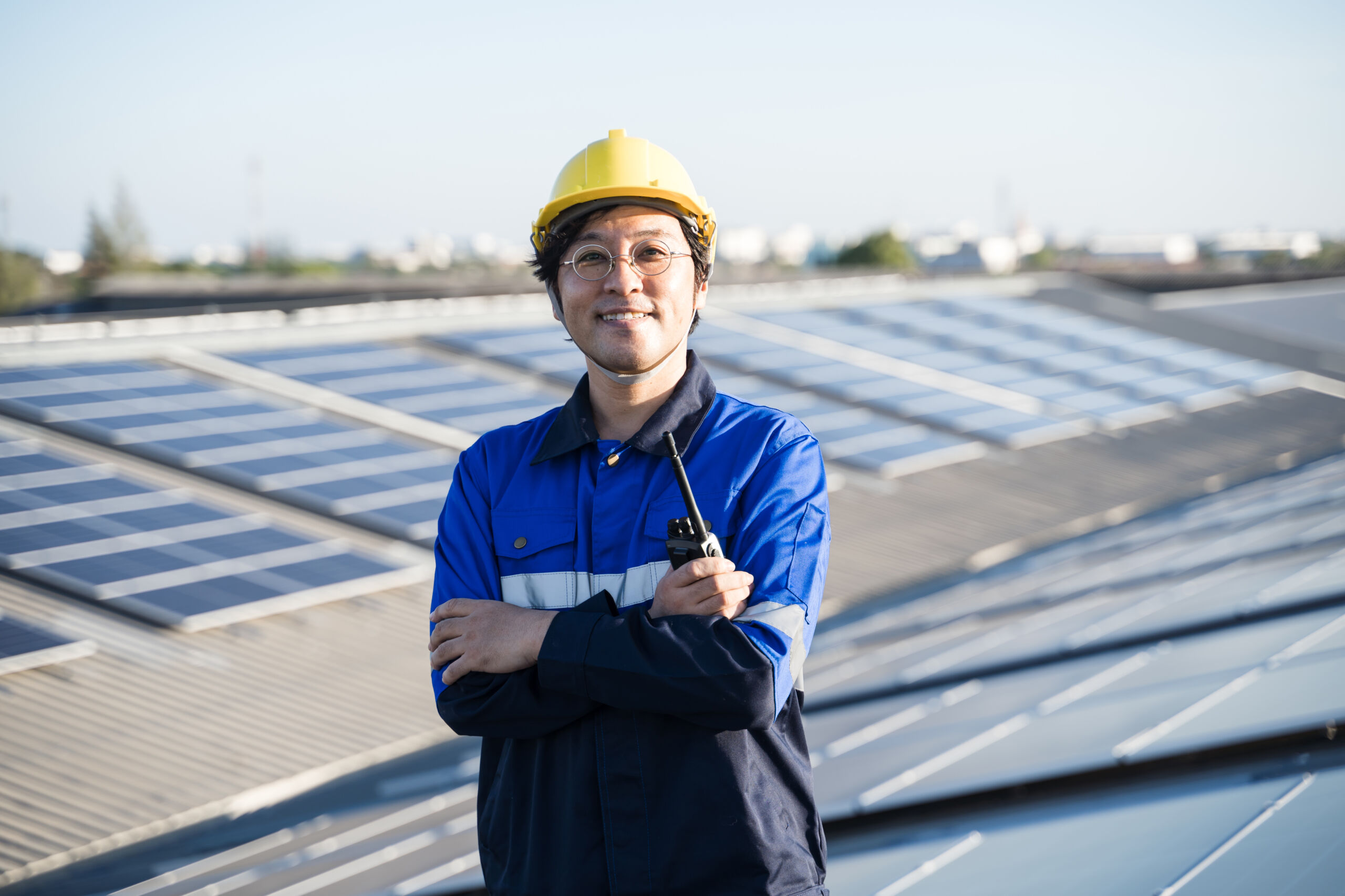 A worker in a blue uniform and yellow hard hat stands with arms folded, holding a walkie-talkie, in front of solar panels on a rooftop.