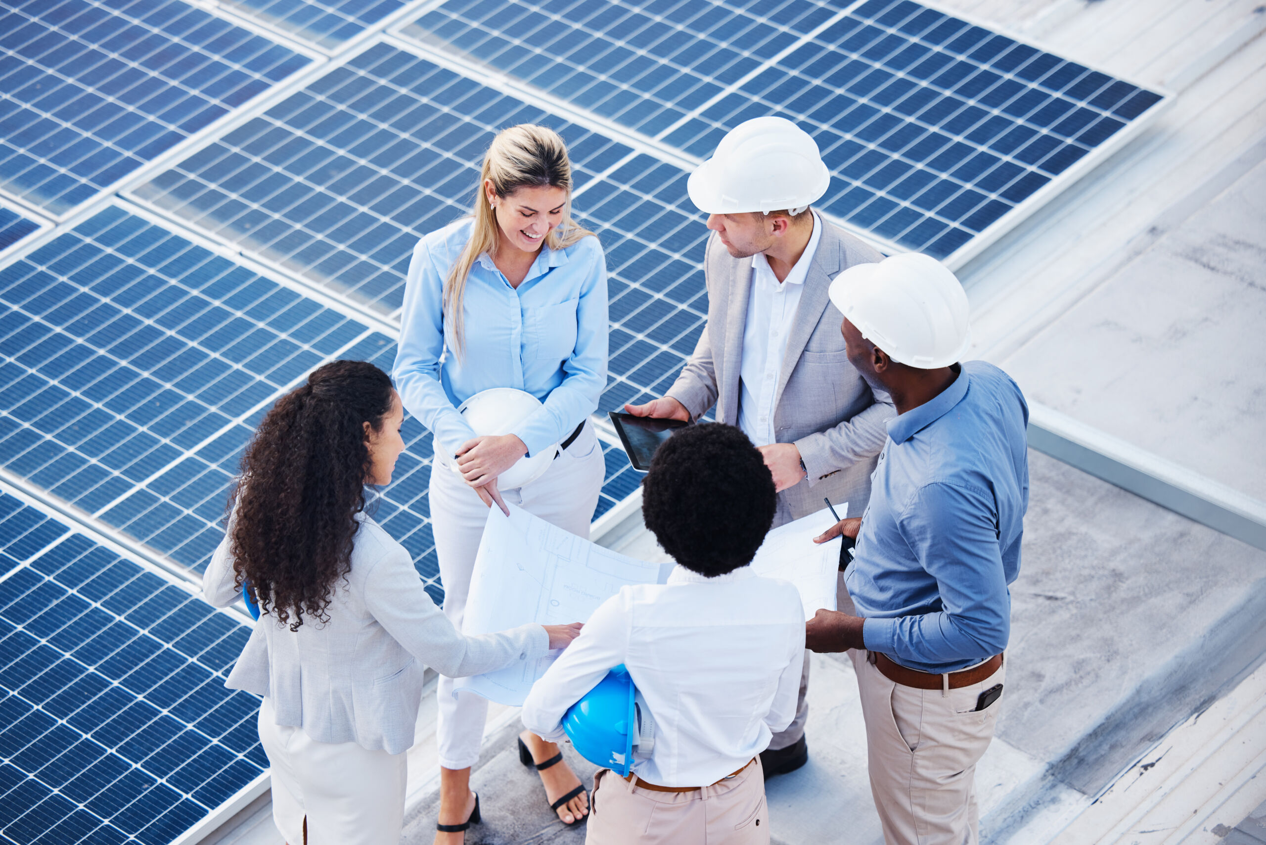 Five people wearing business and safety attire stand on a rooftop with solar panels, reviewing documents and a tablet, appearing to discuss the project.