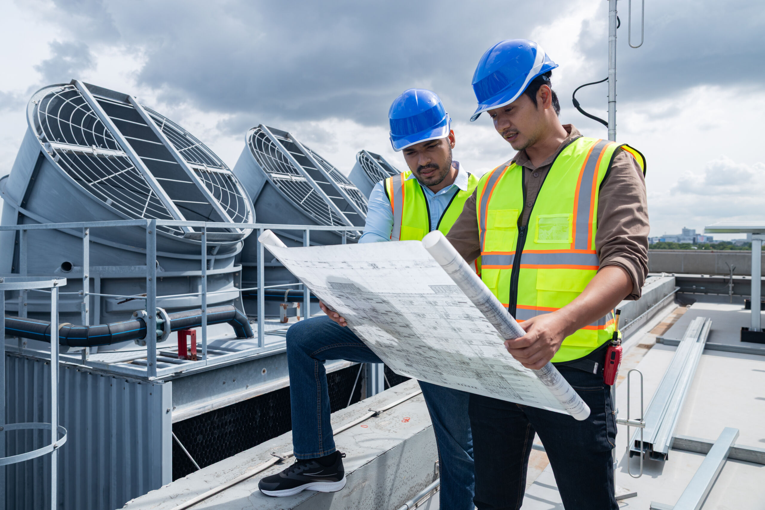 Two engineers wearing safety vests and blue helmets examine plans on a rooftop near large industrial ventilation units under a cloudy sky.