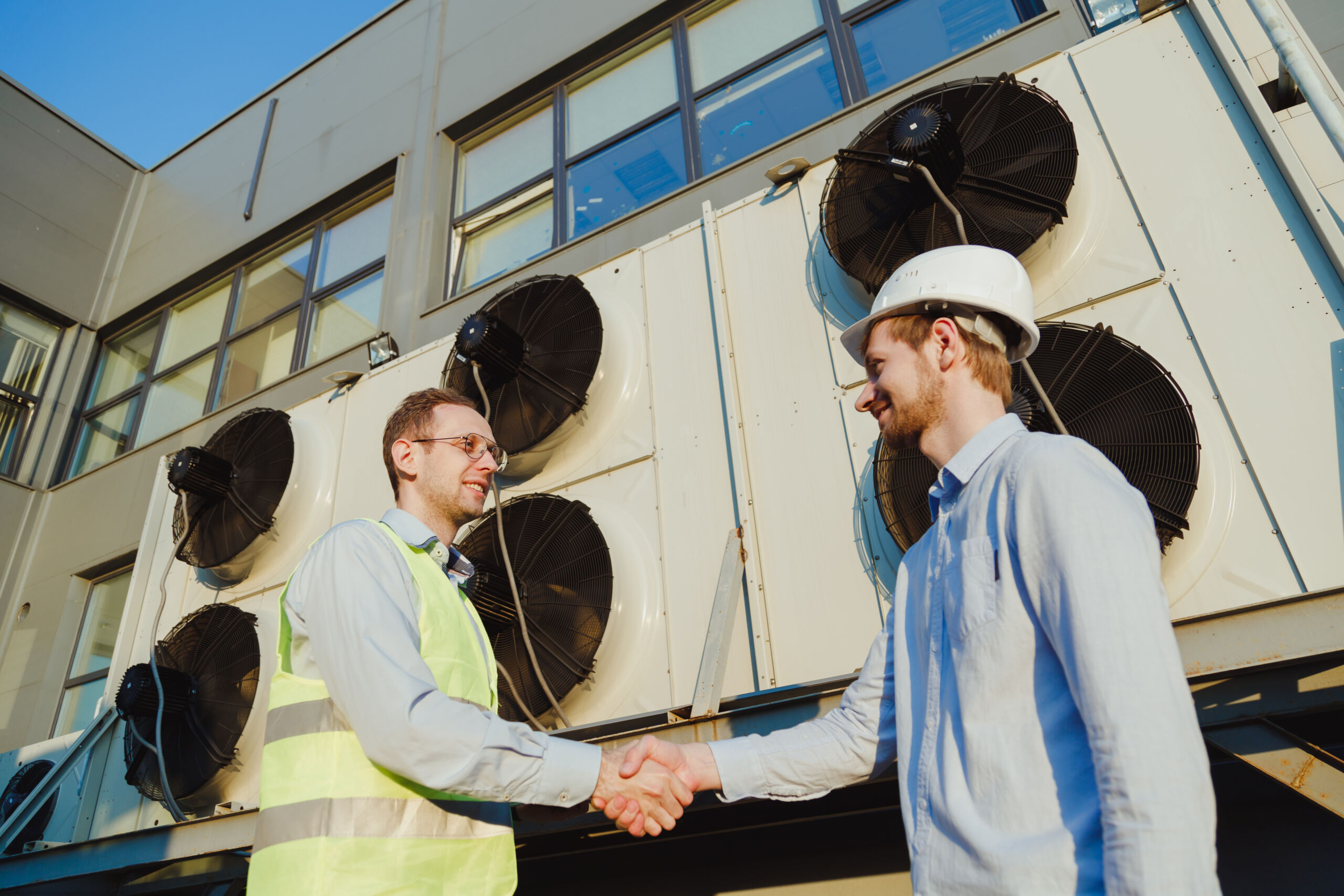 Two men, one in a hi-vis vest and the other in a hard hat, shaking hands in front of large industrial air conditioning units outside a building.