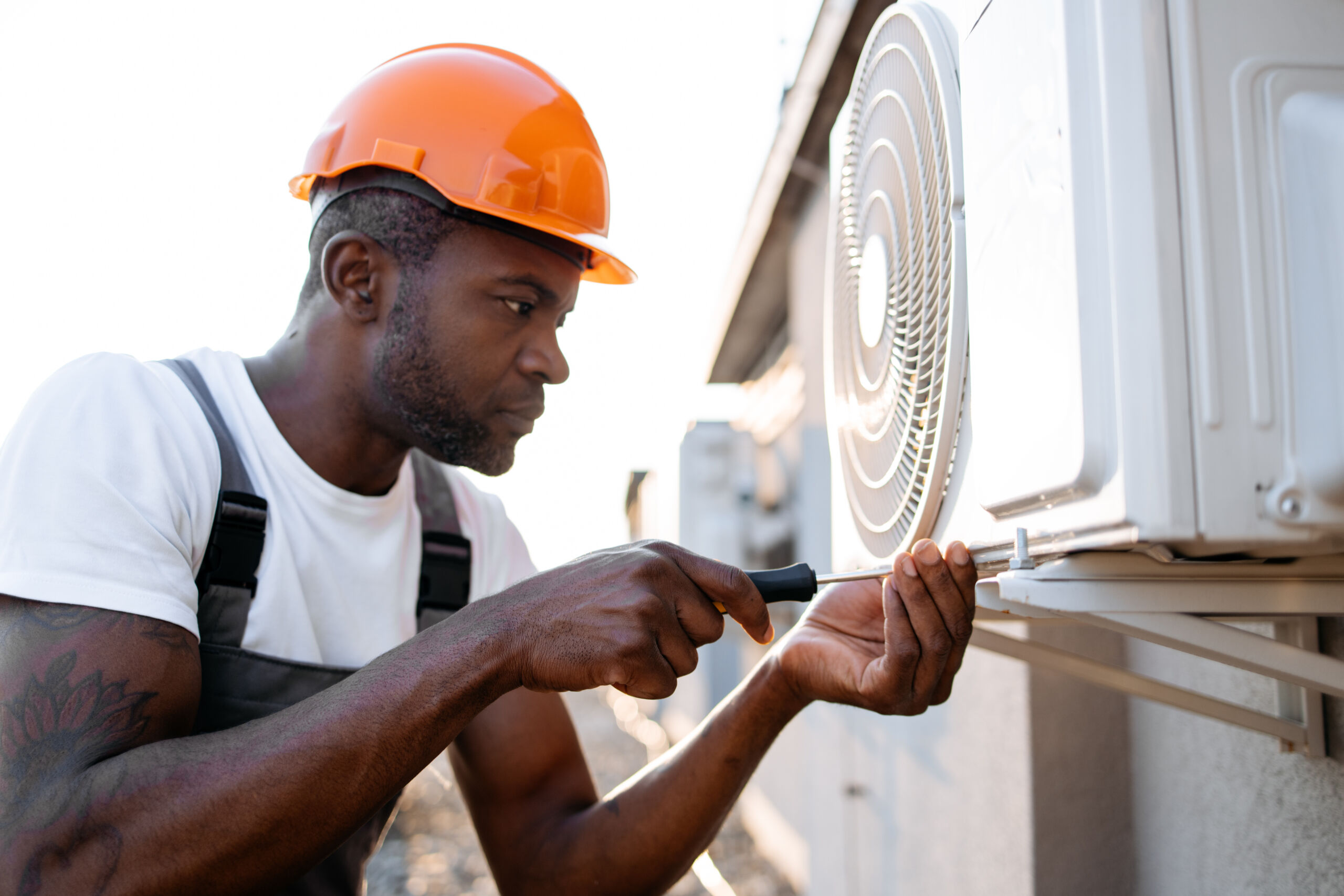 A worker wearing an orange hard hat uses a screwdriver to repair or install an outdoor air conditioning unit mounted on a wall.