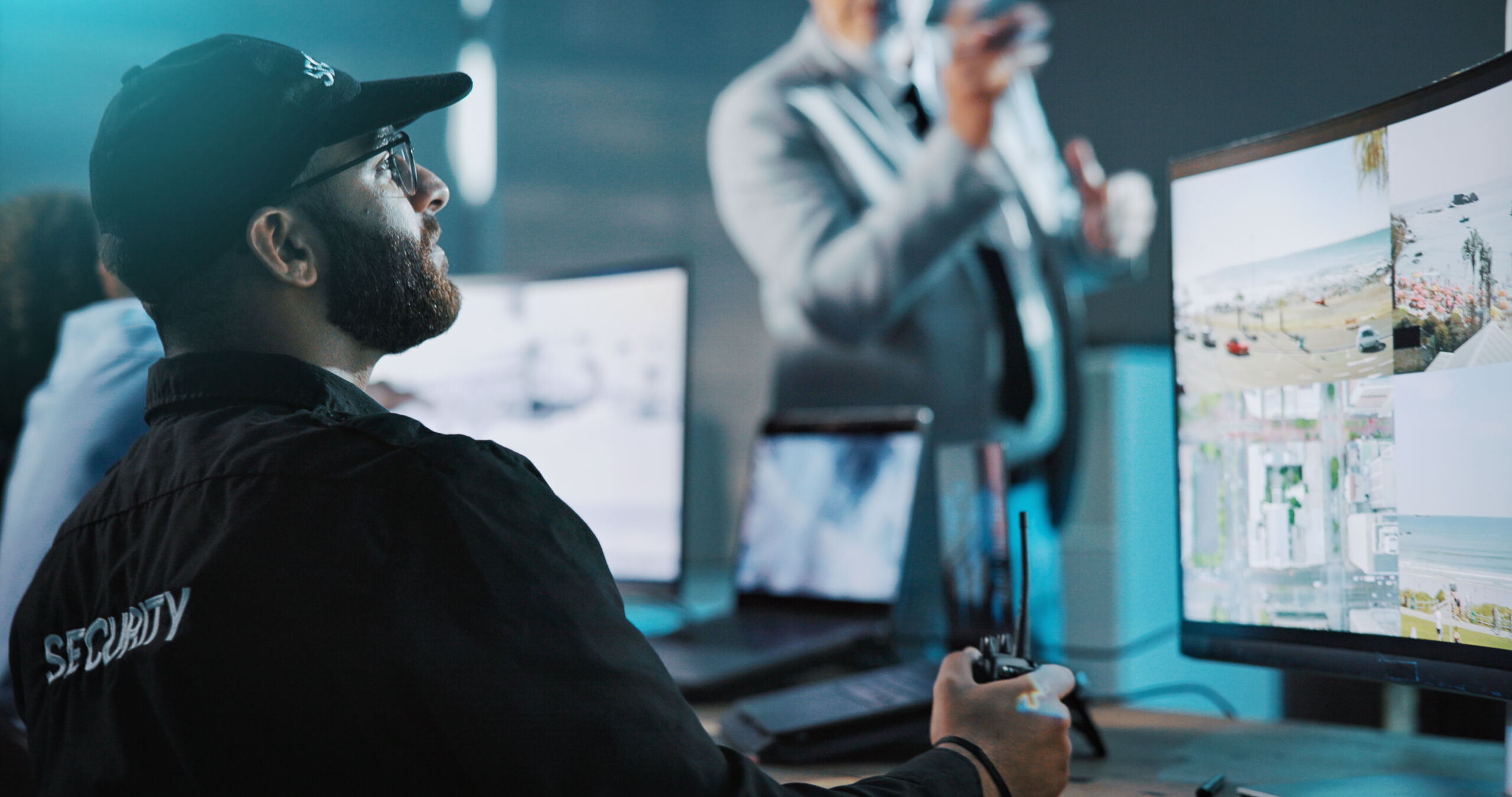 A security officer monitors CCTV footage on multiple screens while holding a two-way radio; another person stands in the blurred background.