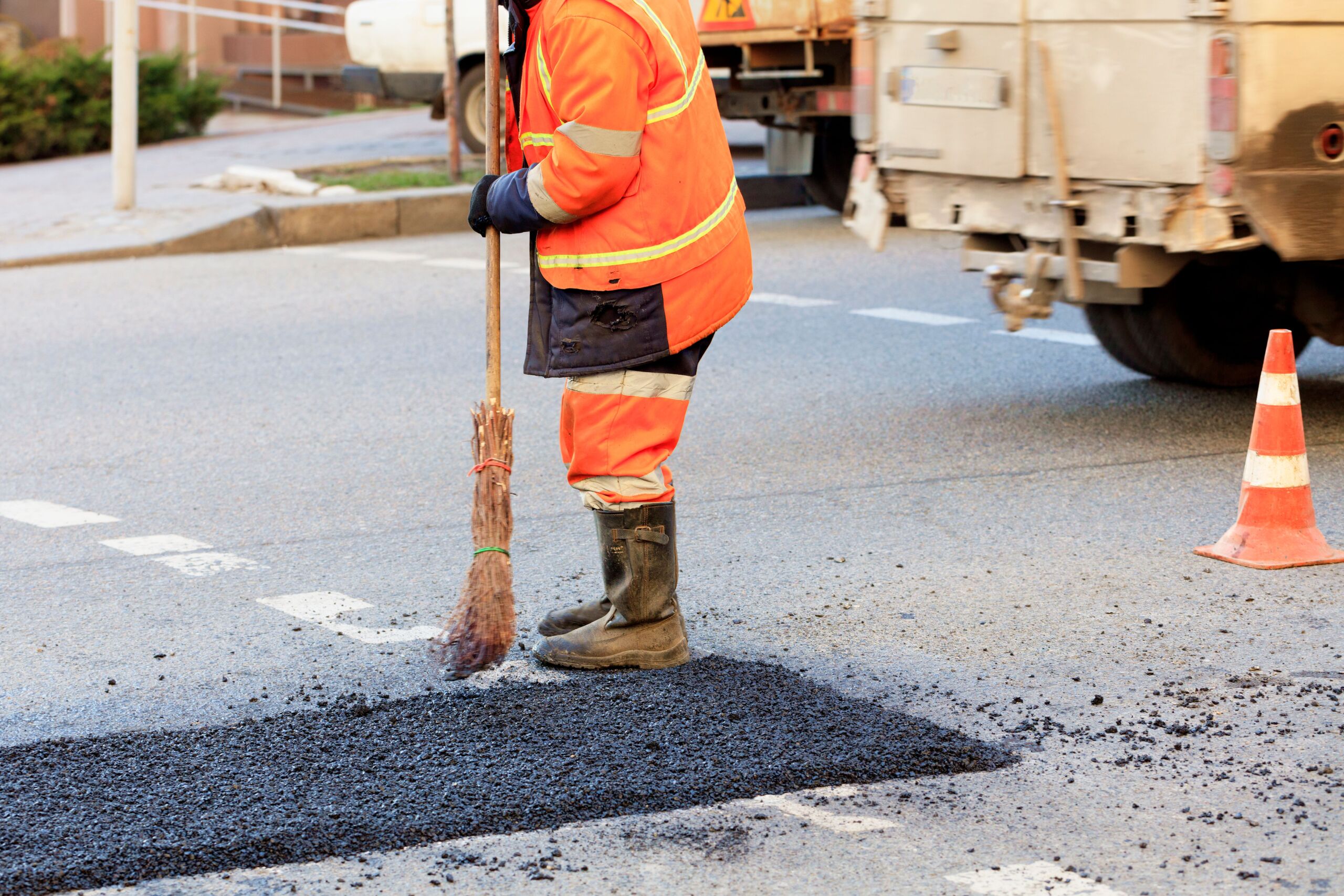 A worker in a reflective orange jacket and boots sweeps debris near freshly laid tarmac on a city street next to a traffic cone and a lorry.