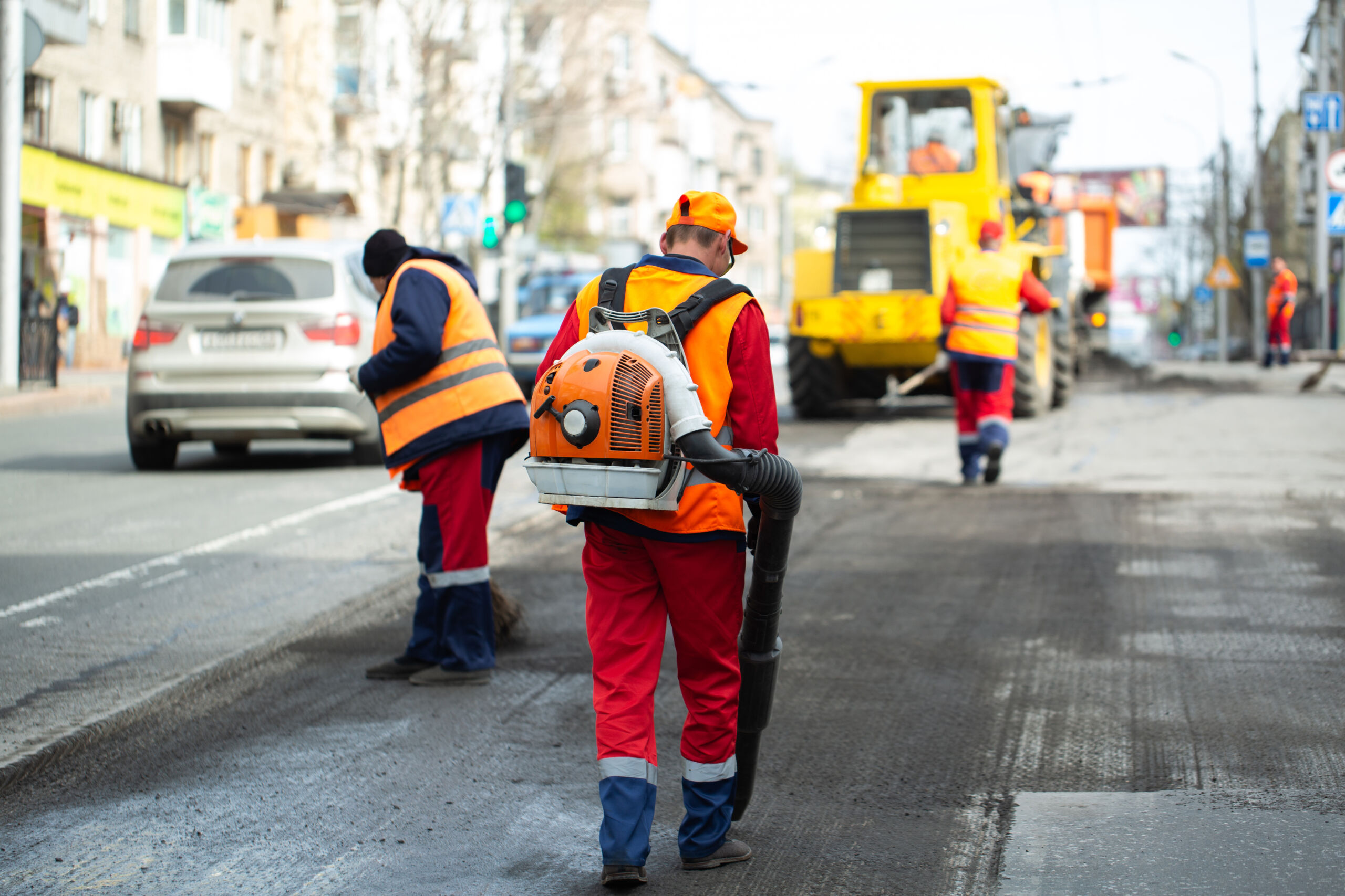 Workers in orange safety vests and uniforms use equipment to clean and repair a city street while cars pass by and heavy machinery operates in the background.