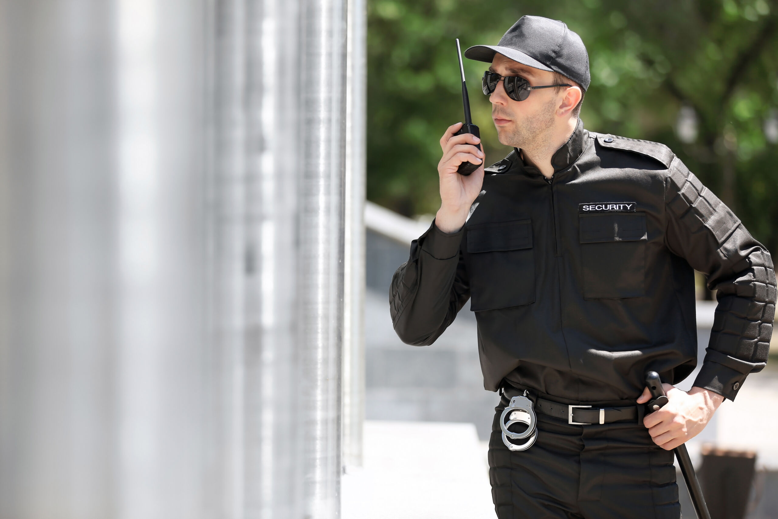 A security guard in a black uniform and cap stands outdoors, holding a walkie-talkie and looking to the side.