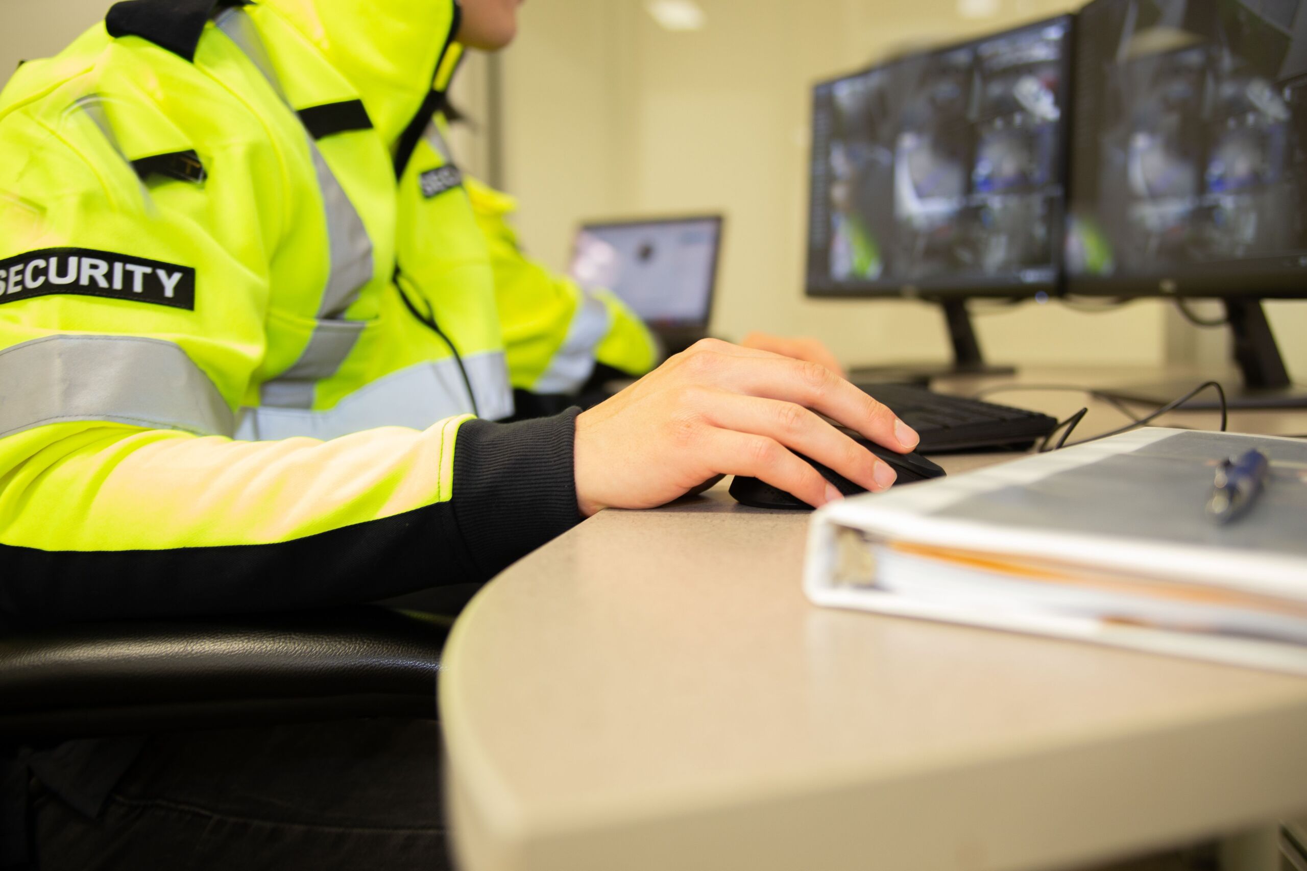 A person in a high-visibility security jacket sits at a desk using a computer mouse, with surveillance monitors and a folder visible on the desk.