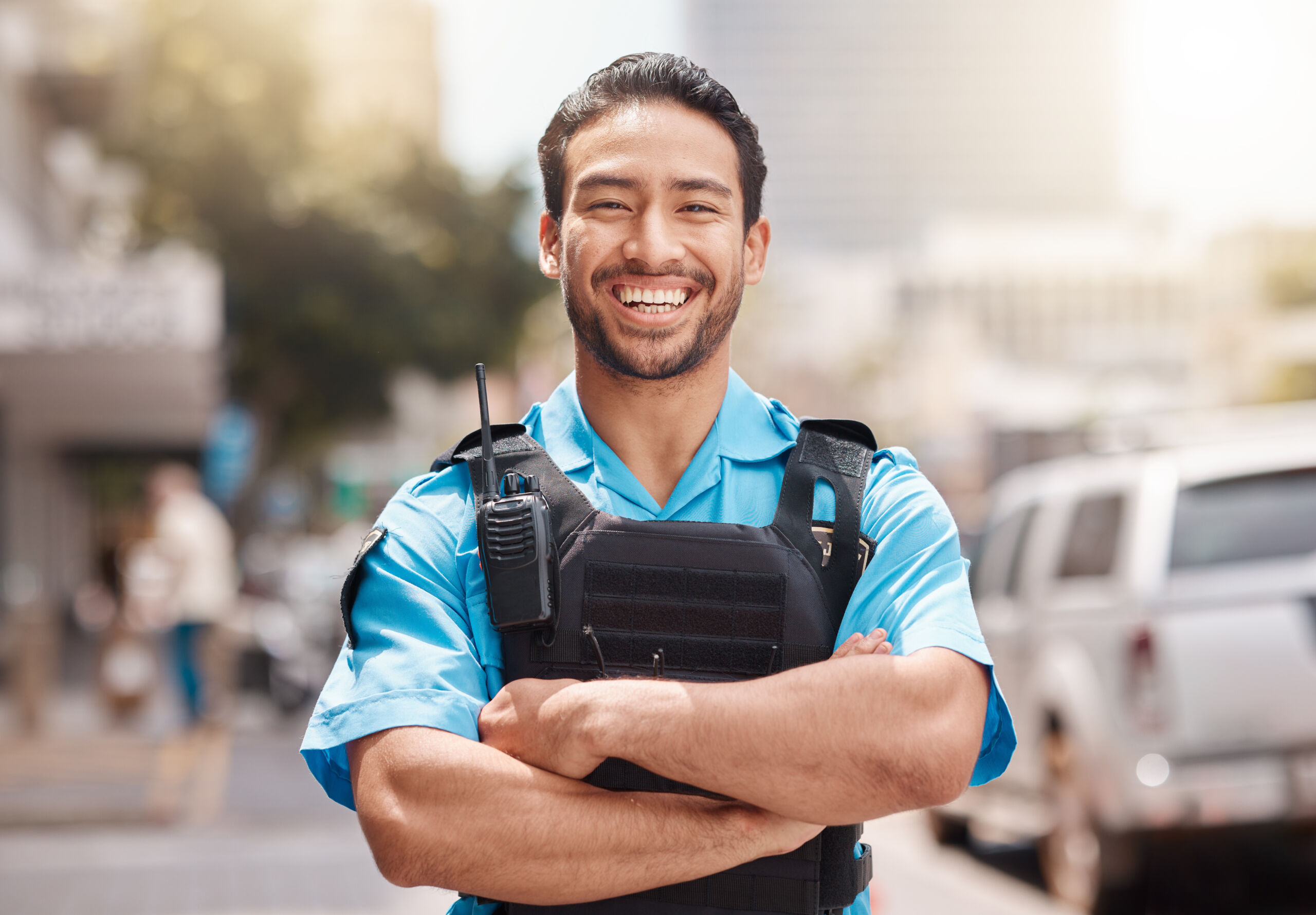 A police officer in uniform stands outdoors with arms folded, smiling, and wearing a radio on a vest. Cars and buildings are visible in the background.