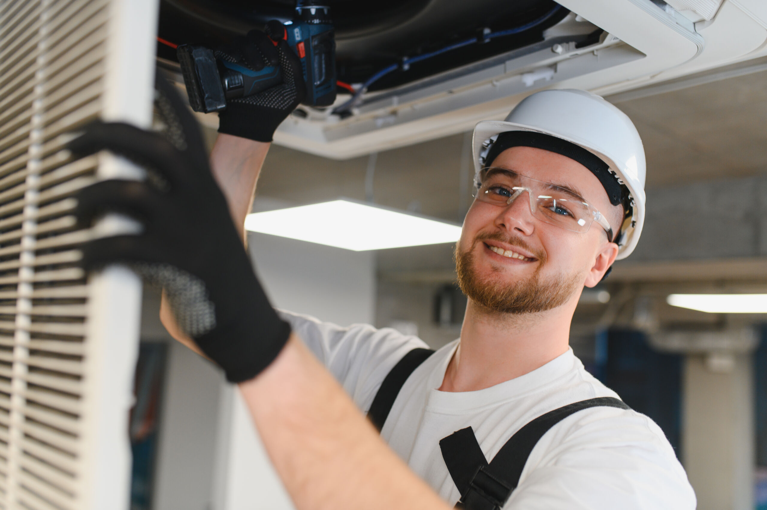 Man wearing a hard hat and safety glasses uses a power drill to install or repair an air conditioning unit in a ceiling.