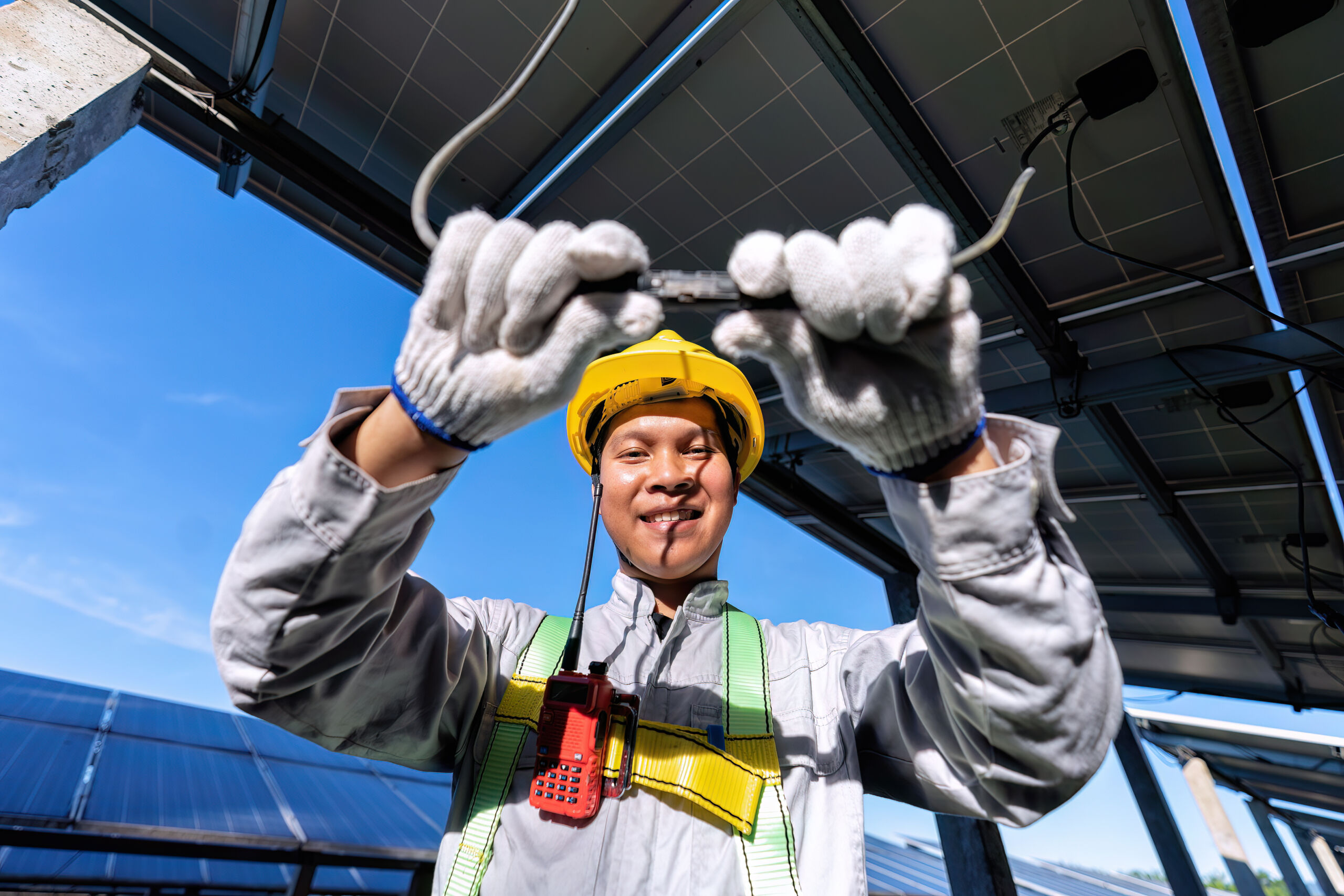 A worker in protective gear and a yellow helmet holds up electrical wires under solar panels, with a blue sky in the background.