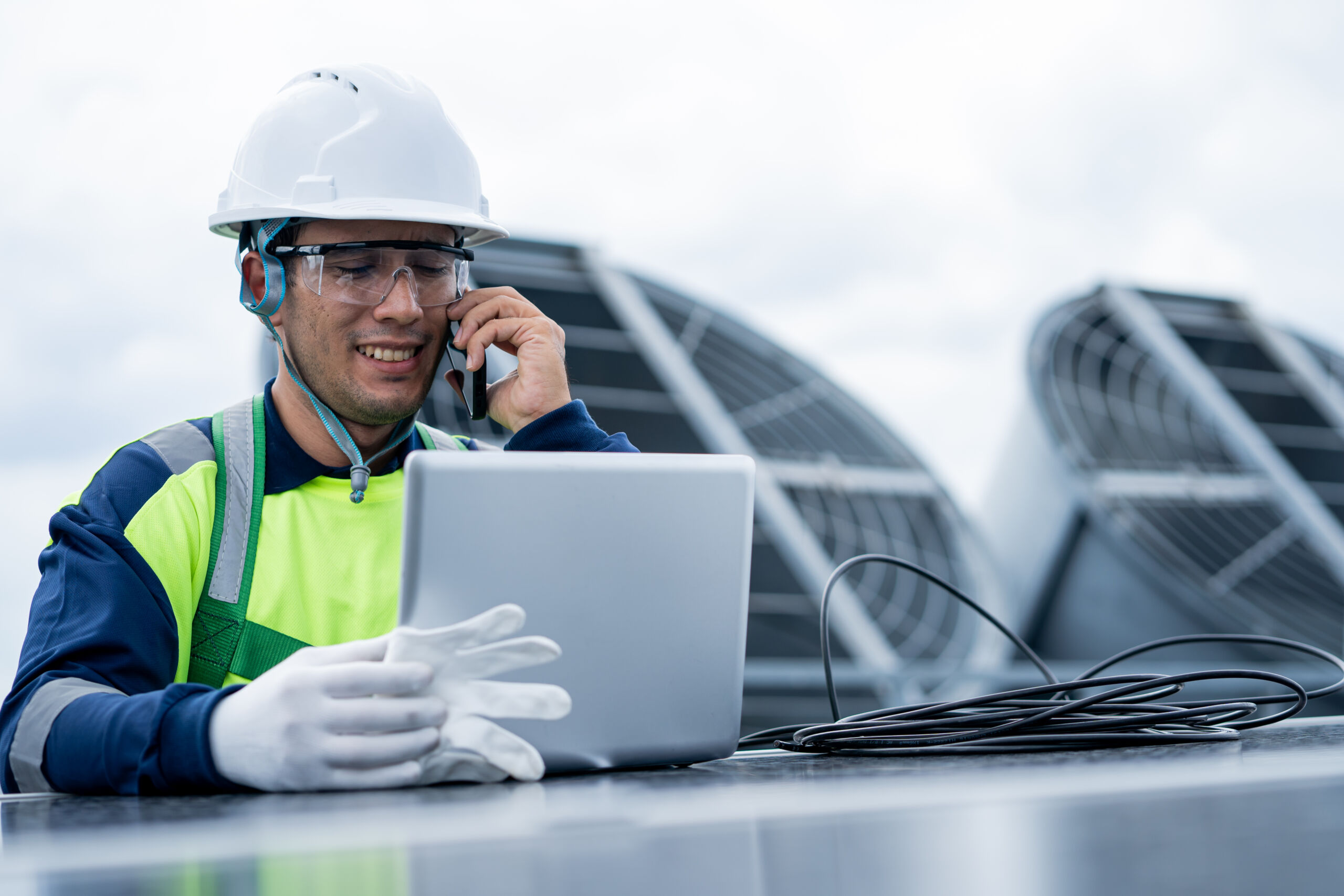 A worker in safety gear uses a laptop and talks on the phone outdoors near industrial ventilation units.