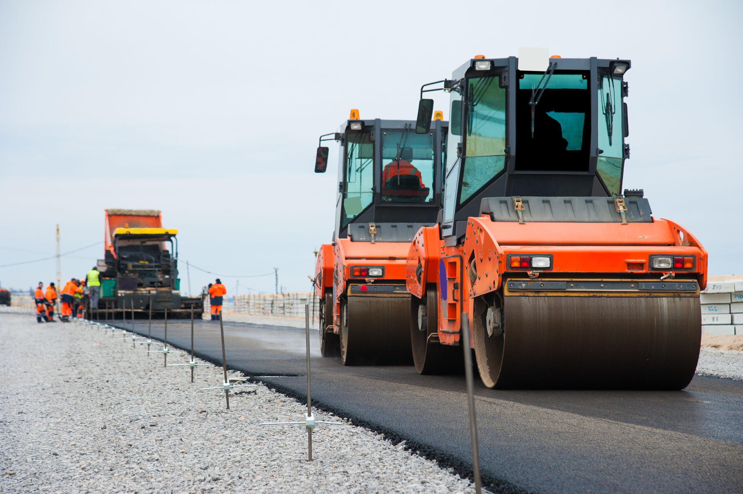 Construction workers and heavy machinery lay and smooth tarmac on a new road, with two steamrollers in the foreground and more workers and equipment in the background.